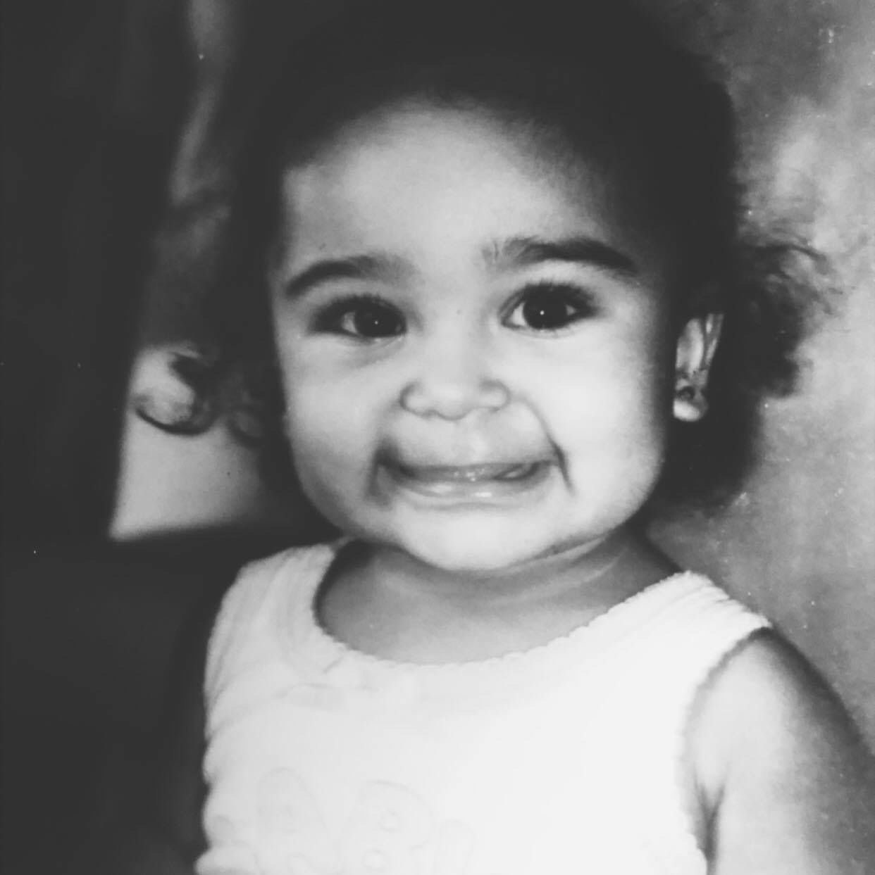 Portrait of a smiling woman with long wavy hair wearing a blue sweatshirt and gold hoop earrings against a gray background.