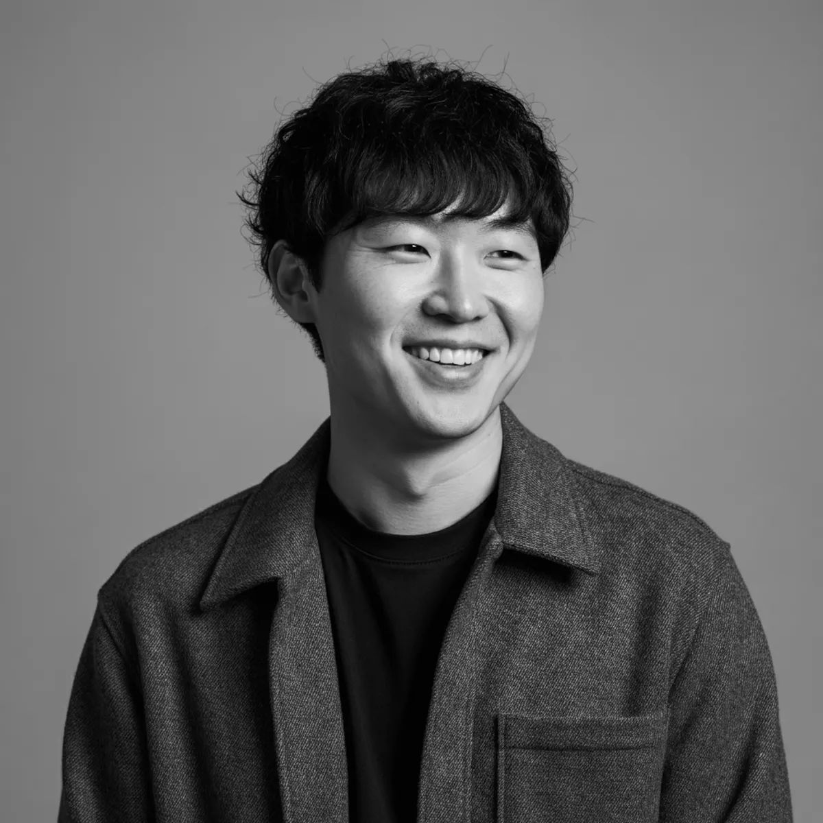 Smiling man with wavy black hair in a gray jacket and black shirt, set against a neutral gray background.