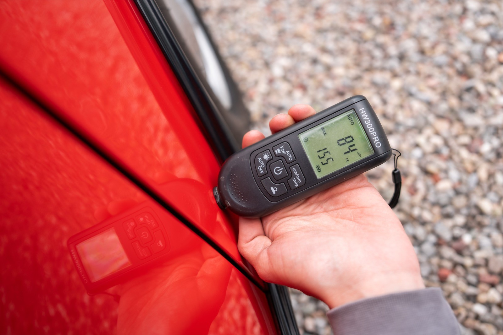 A hand holds a digital paint thickness gauge against a red car door. The display shows a numeric reading.