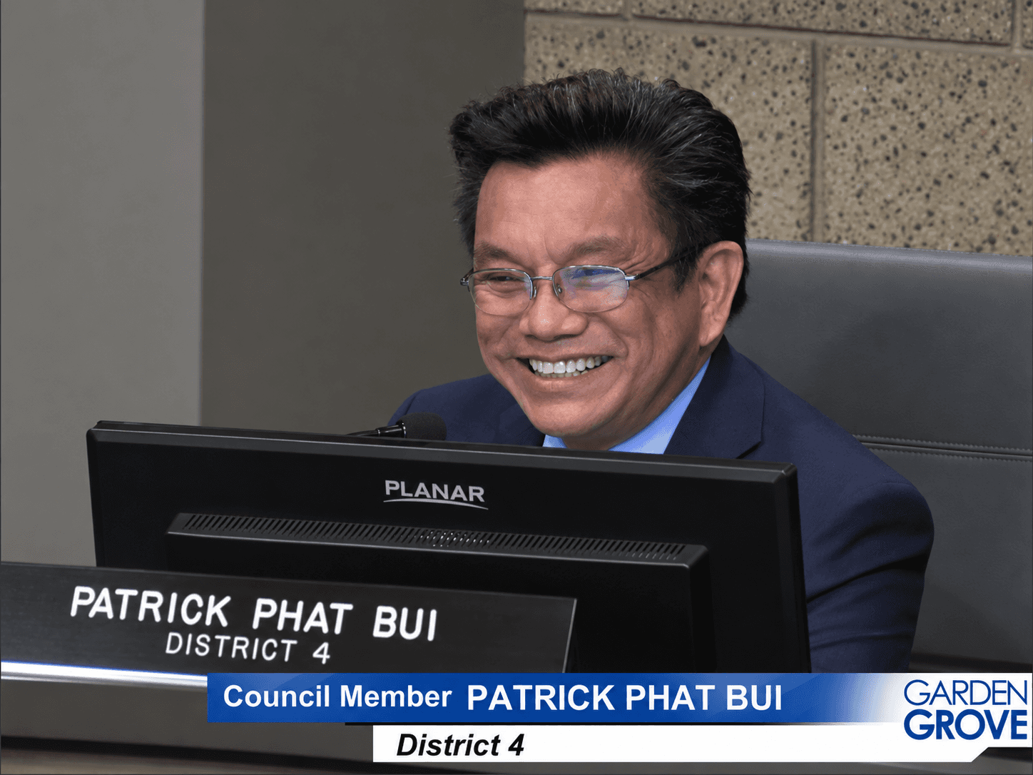 Phát Bùi, mặc com-lê xanh đậm và áo sơ mi xanh nhạt, đang cười khi ngồi sau một màn hình máy tính và một micrô. Bảng tên trước mặt ông ghi Patrick Phat Bui, District 4 (Địa Hạt 4), và dòng chú thích ở cuối ảnh ghi Council Member Patrick Phat Bui, District 4 (Nghị Viên Patrick Phát Bùi, Địa Hạt 4), bên cạnh biểu trưng Garden Grove.