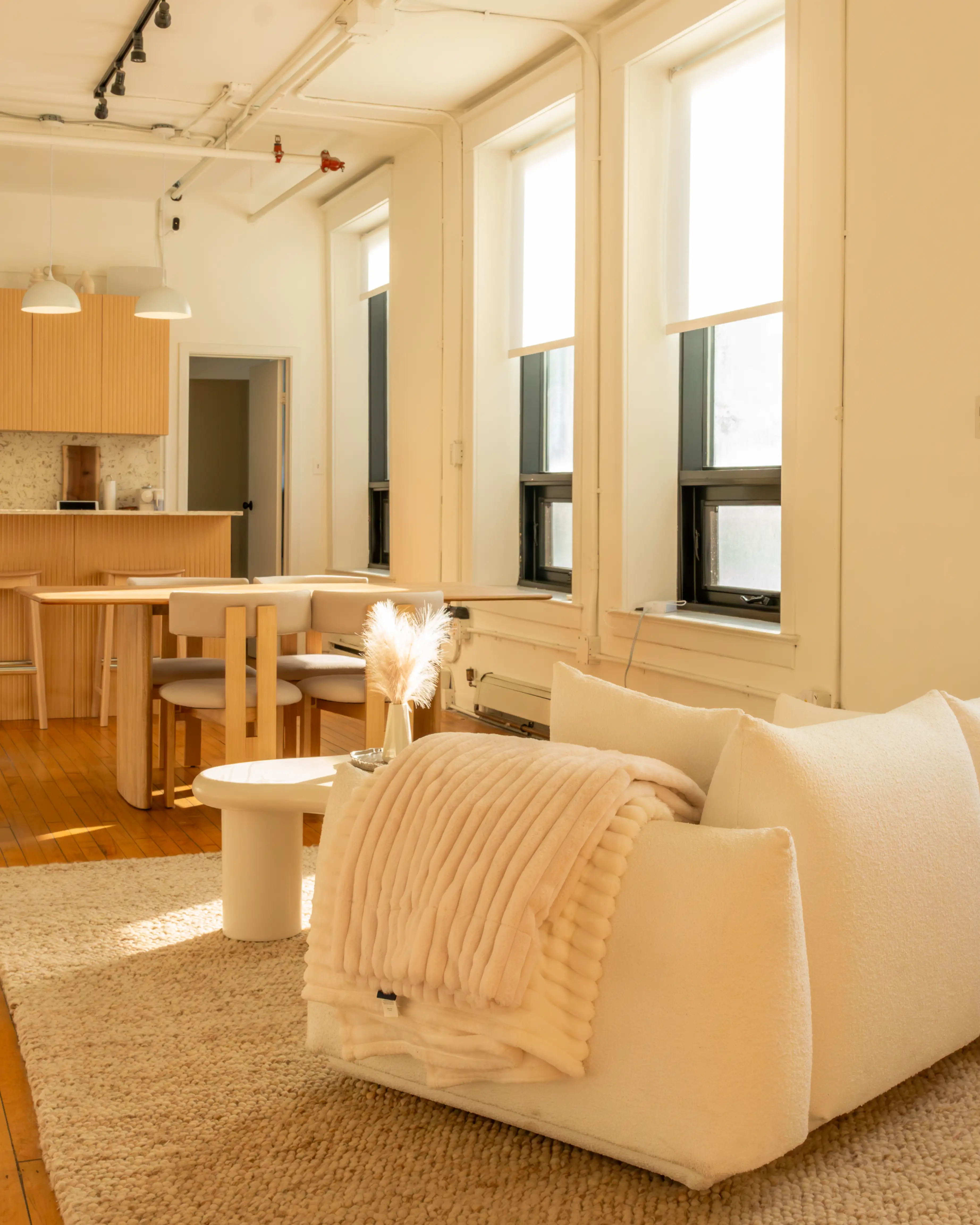 Bright and cozy corner of Node Studio featuring a plush white couch with textured throws, natural light streaming through large windows, hardwood floors, and a view of the semi-equipped kitchen with minimalist wooden furniture
