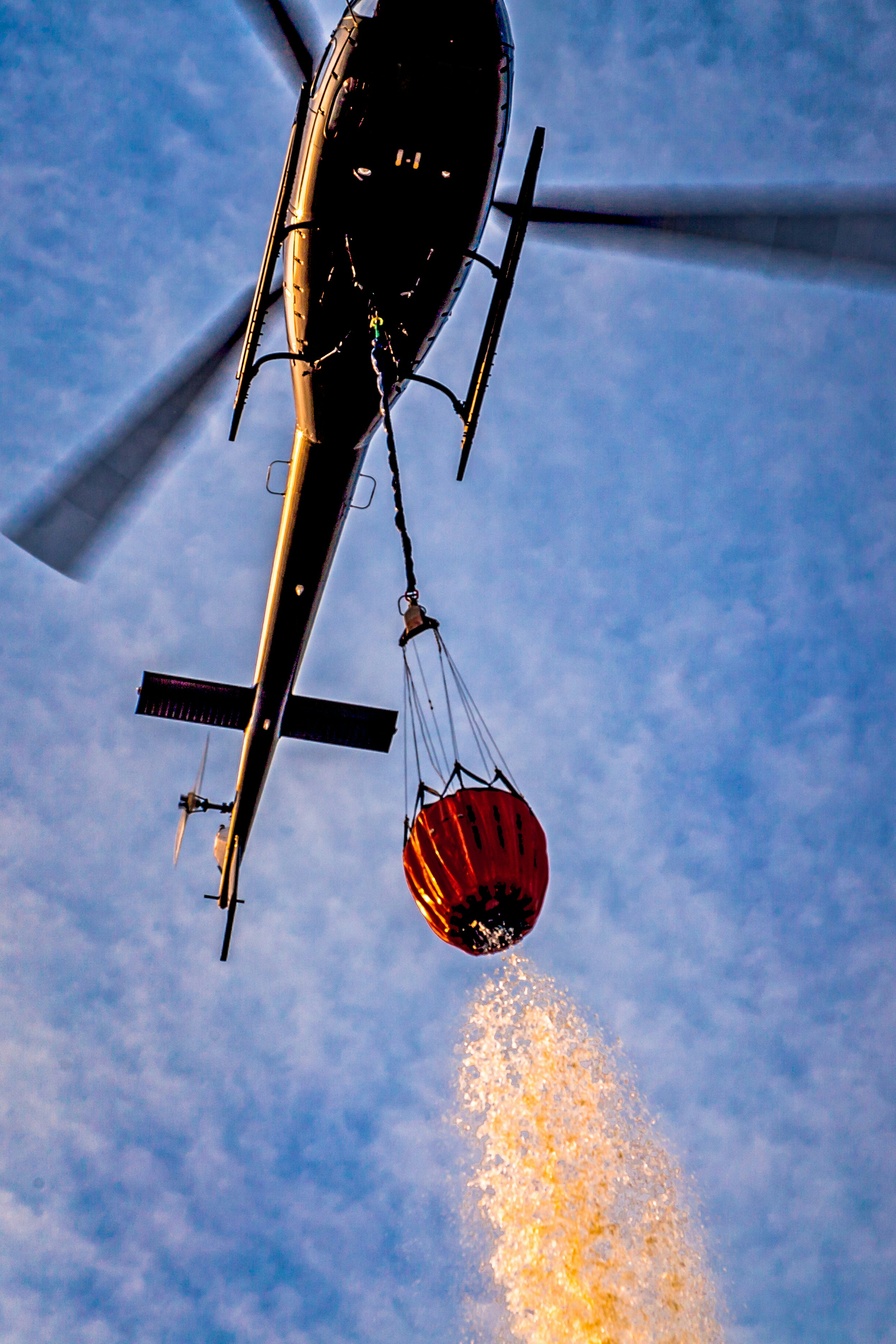 Helicopter dropping water from a bucket onto a wildfire from below, against a blue sky