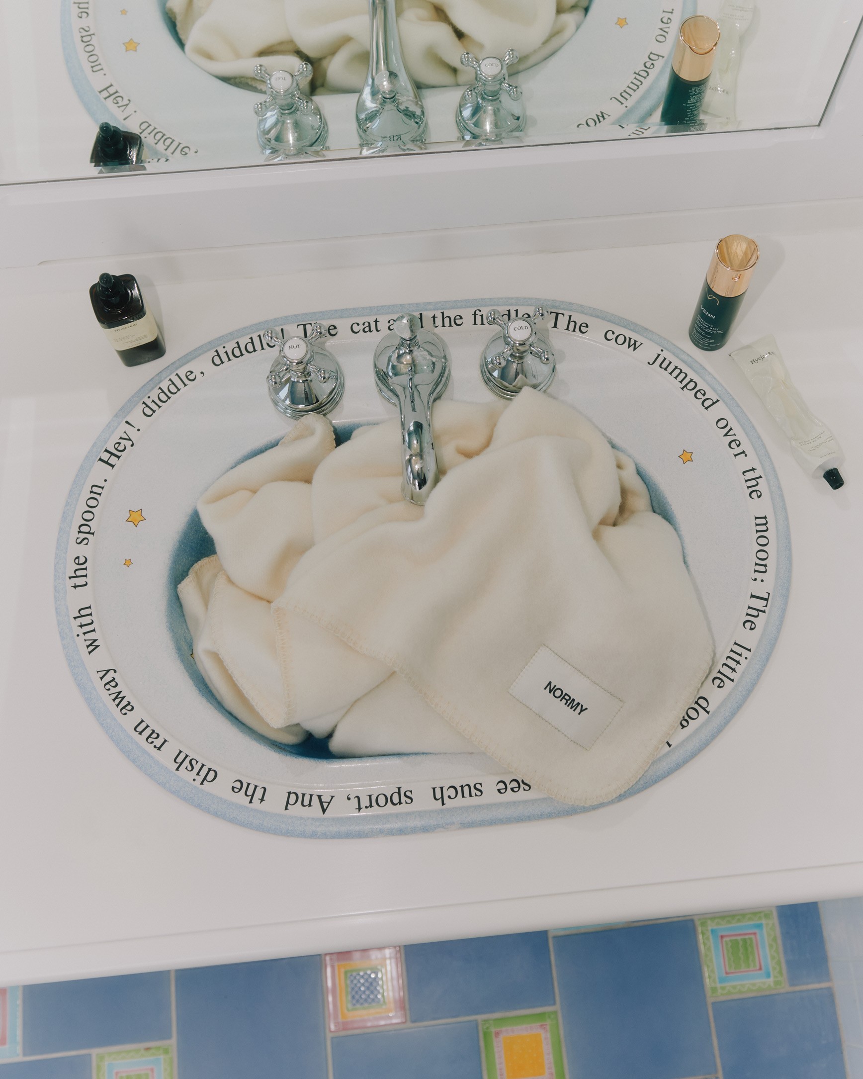 A soft cream-colored Normy blanket with a visible brand label is draped inside a decorative bathroom sink. The scene also includes bath products around the sink and a mirror reflecting the cozy setup, creating a playful and inviting atmosphere.