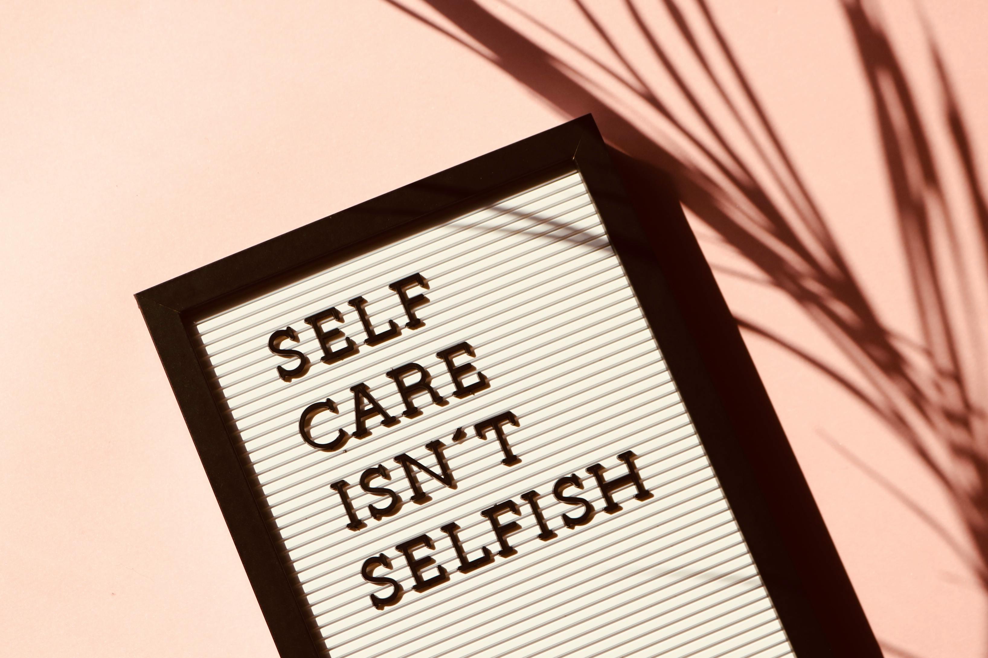 Close-up photograph of a message board with the phrase 'SELF CARE ISN'T SELFISH' in bold black letters. The board is framed in black, set against a pink background with soft shadows of plant leaves cast across the surface, creating a warm and reflective mood