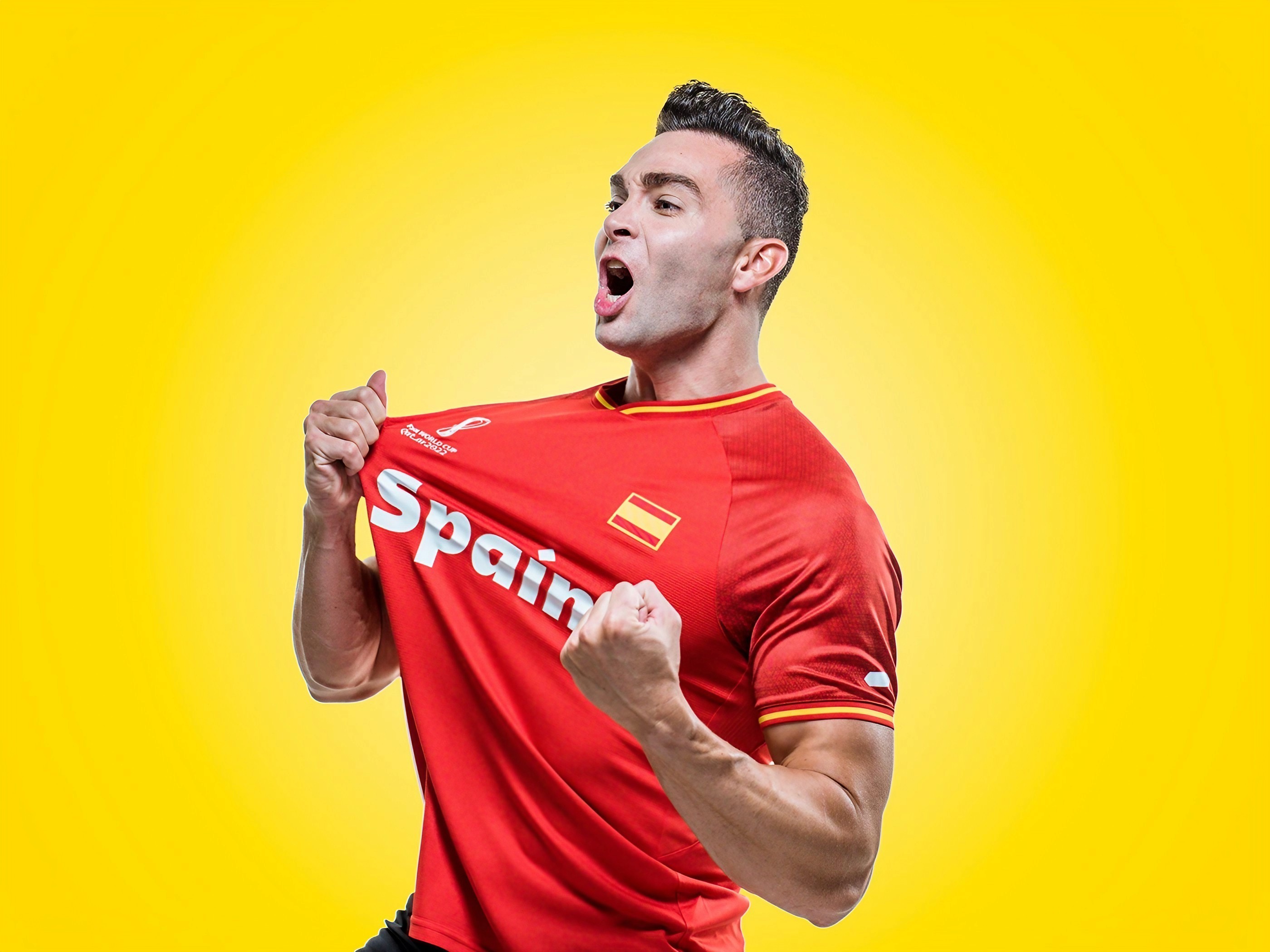 A football fan roaring in celebration, tugging at his red Spain jersey against a vivid yellow background.