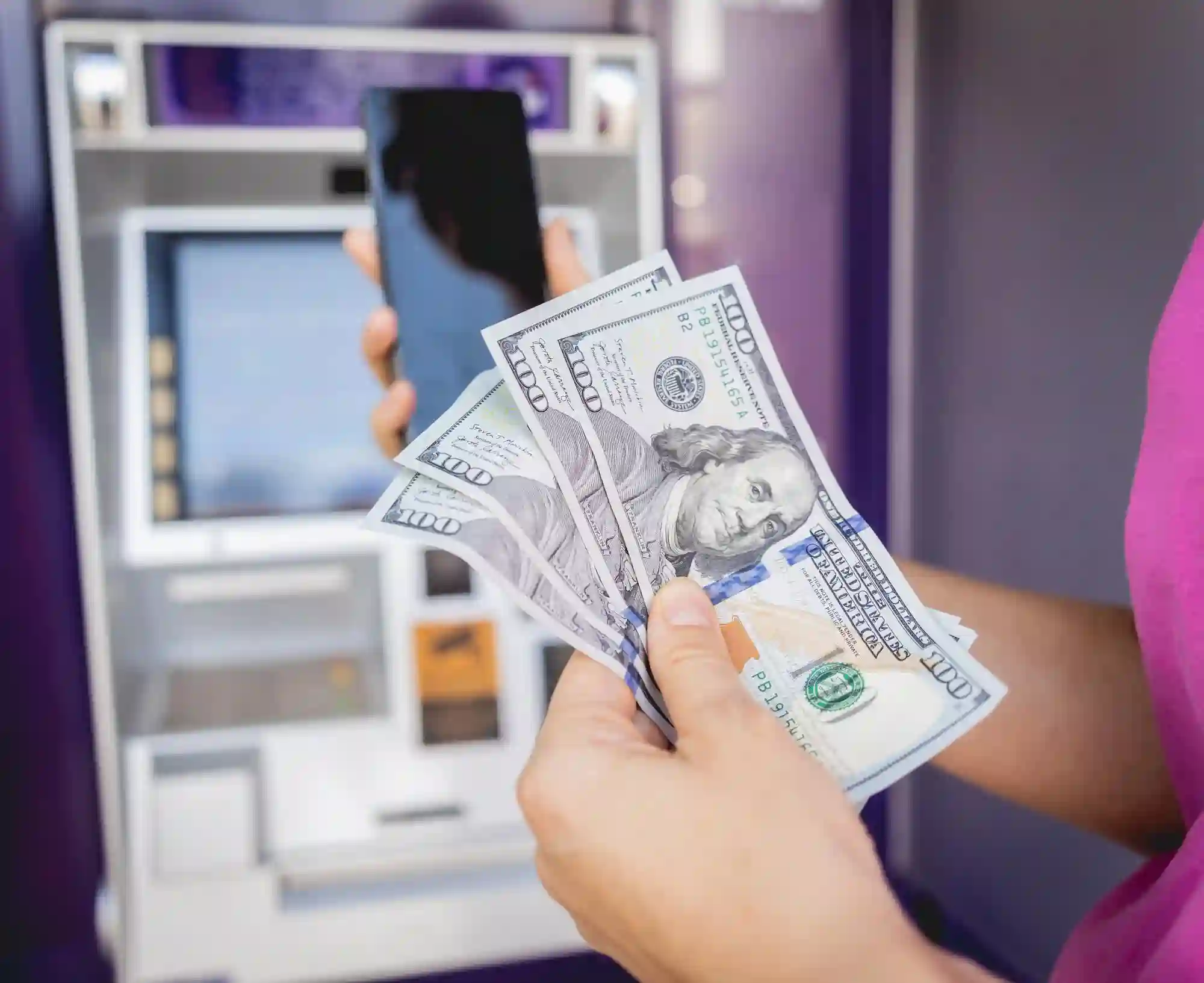 Hands holding US dollars and a smartphone near an ATM.