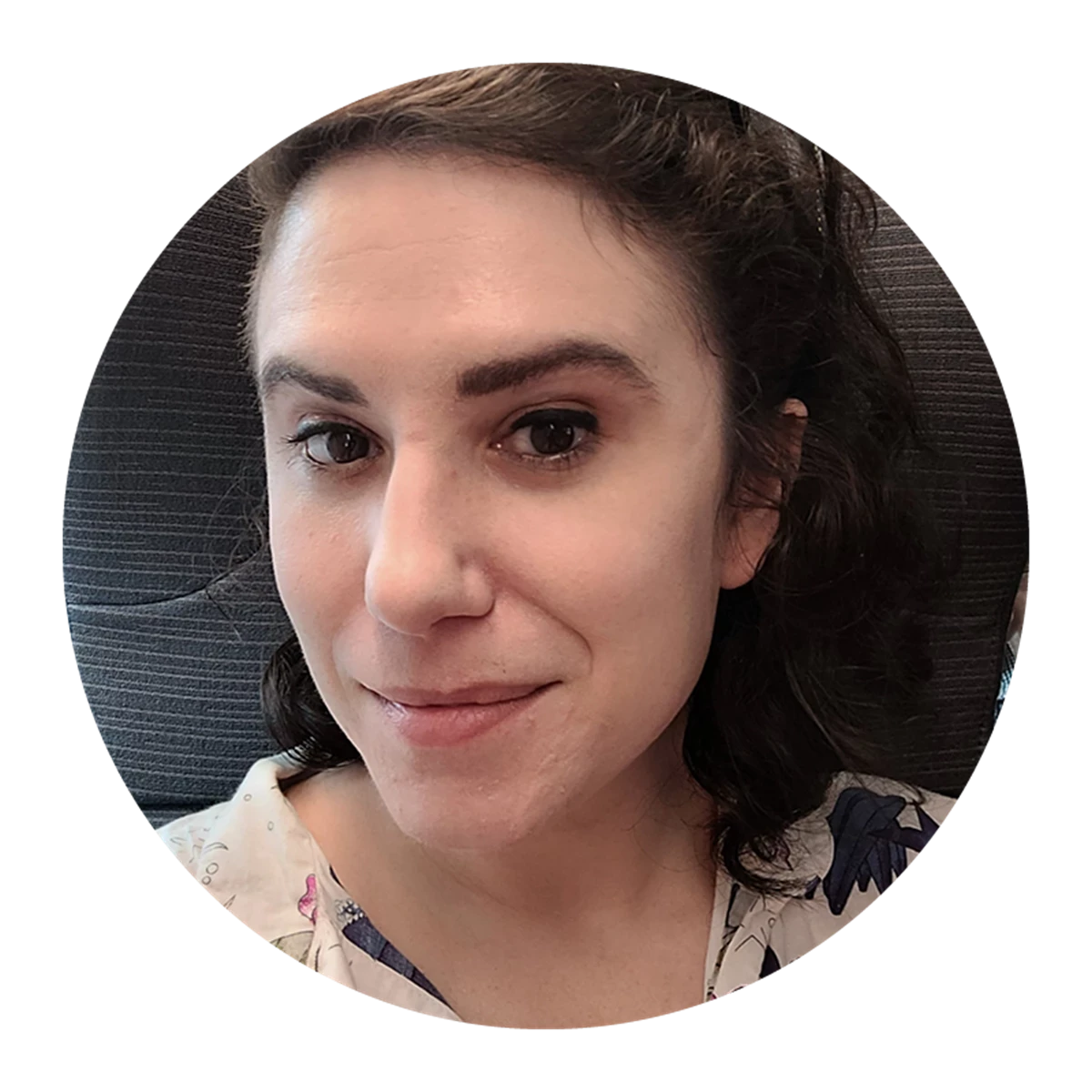 Person with dark curly hair wearing a light top with a floral pattern, seated against a dark fabric background.