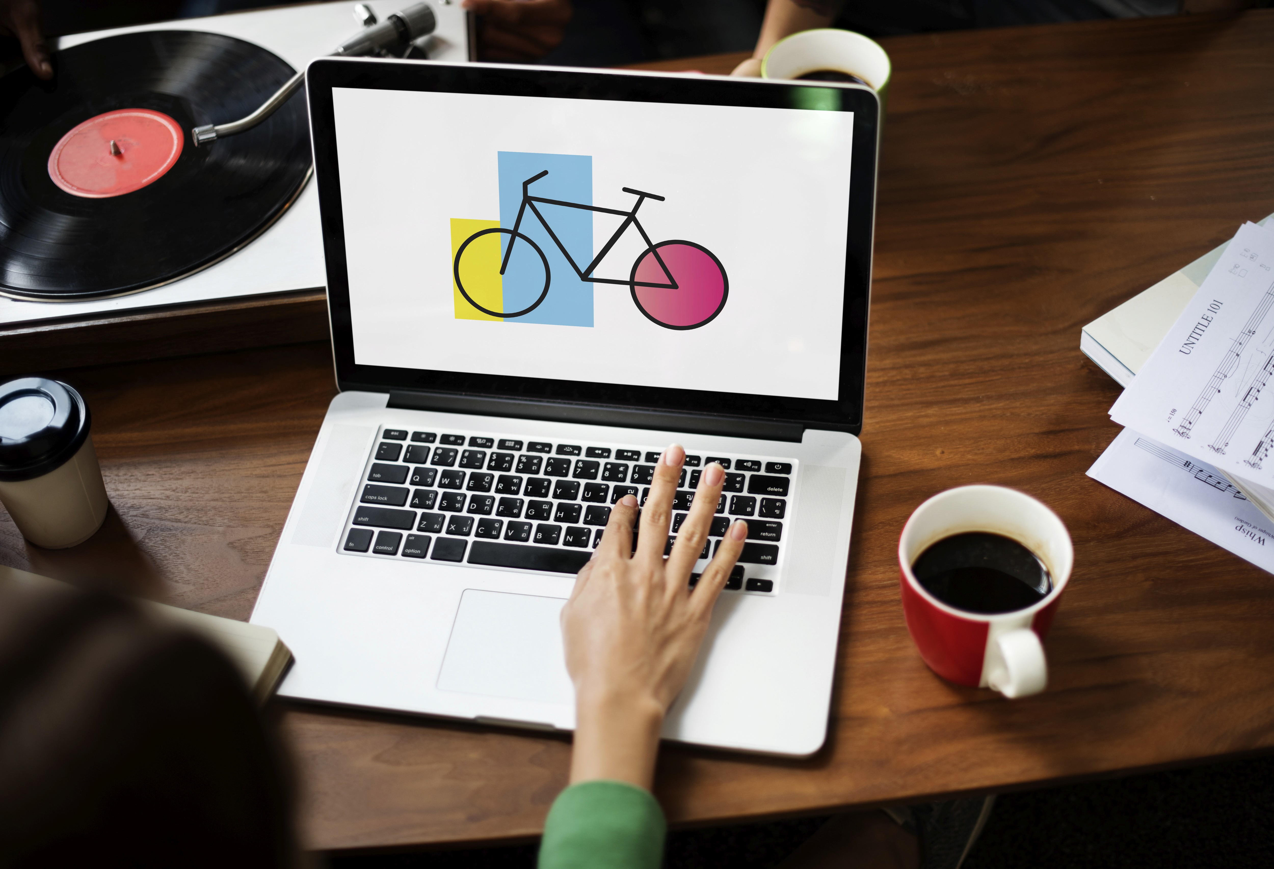 Woman using laptop with colorful geometric bicycle graphic on screen at creative workspace with coffee and vinyl record player