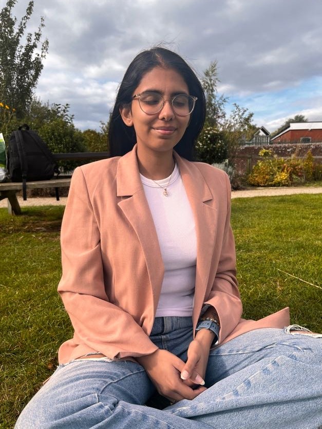 Vrinda in black and white striped top with hands folded near waist, standing against a backdrop of plants, introducing her therapeutic approach.