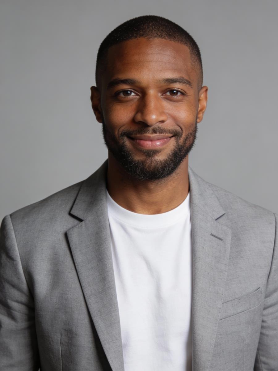 A man with a welcoming smile wears a light gray blazer over a white shirt against a plain gray background, exuding a professional and friendly demeanor.