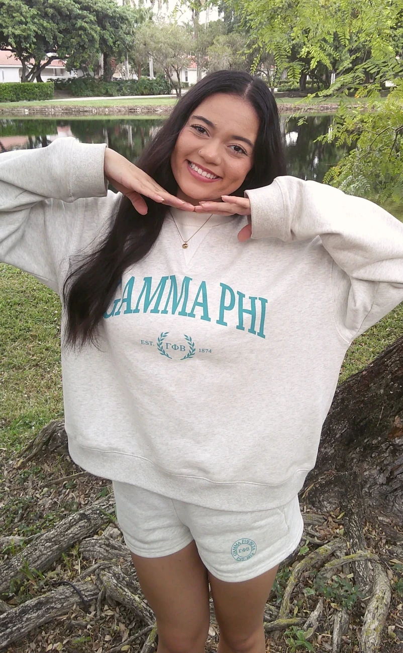 person posing by a lakeside wearing a light sweatshirt reading “GAMMA PHI” and matching shorts