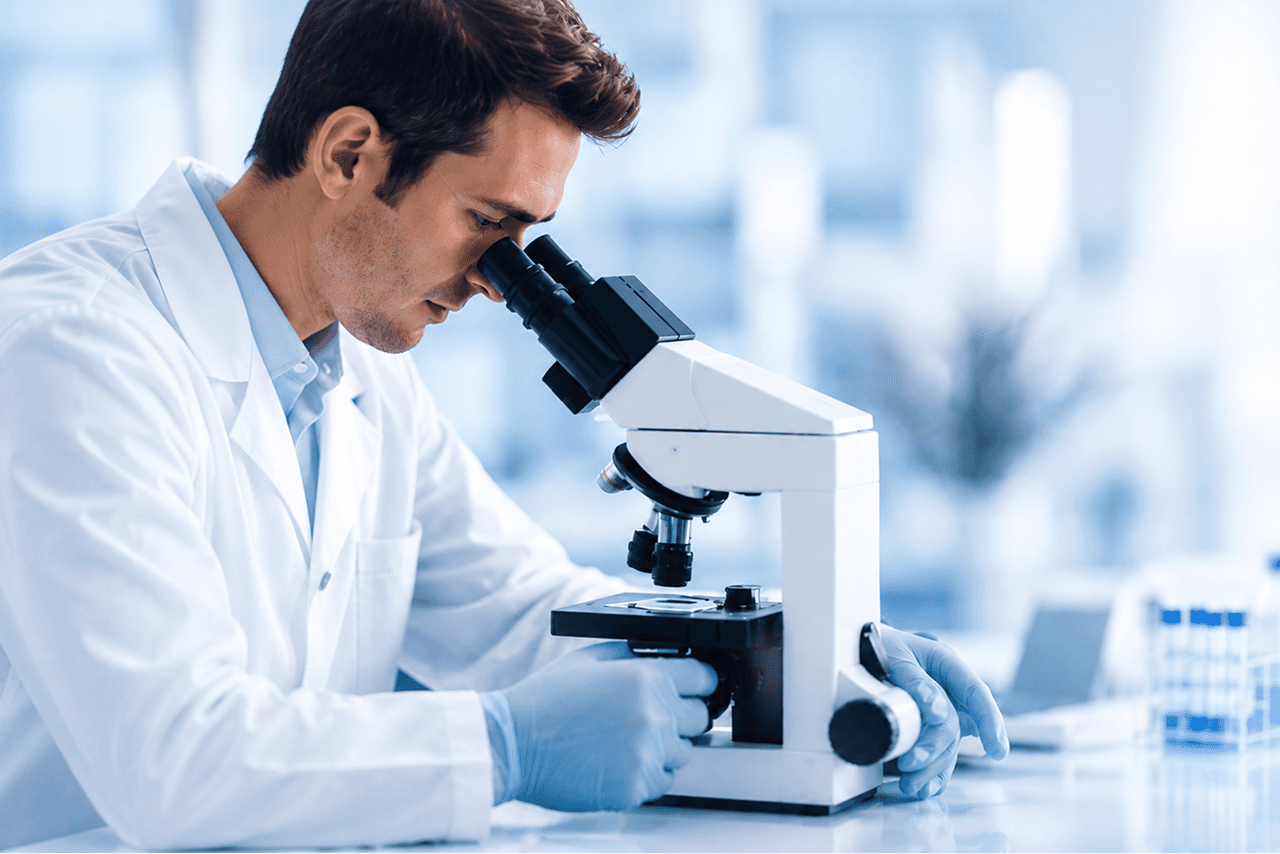 Fehér köpenyes kutató mikroszkóppal vizsgál mintát egy világos laboratóriumban.