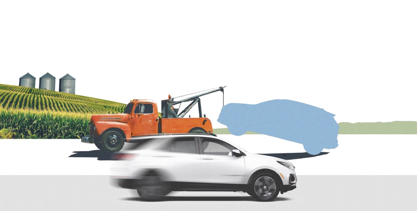 White electric car driving past vintage orange tow truck near farm silos.