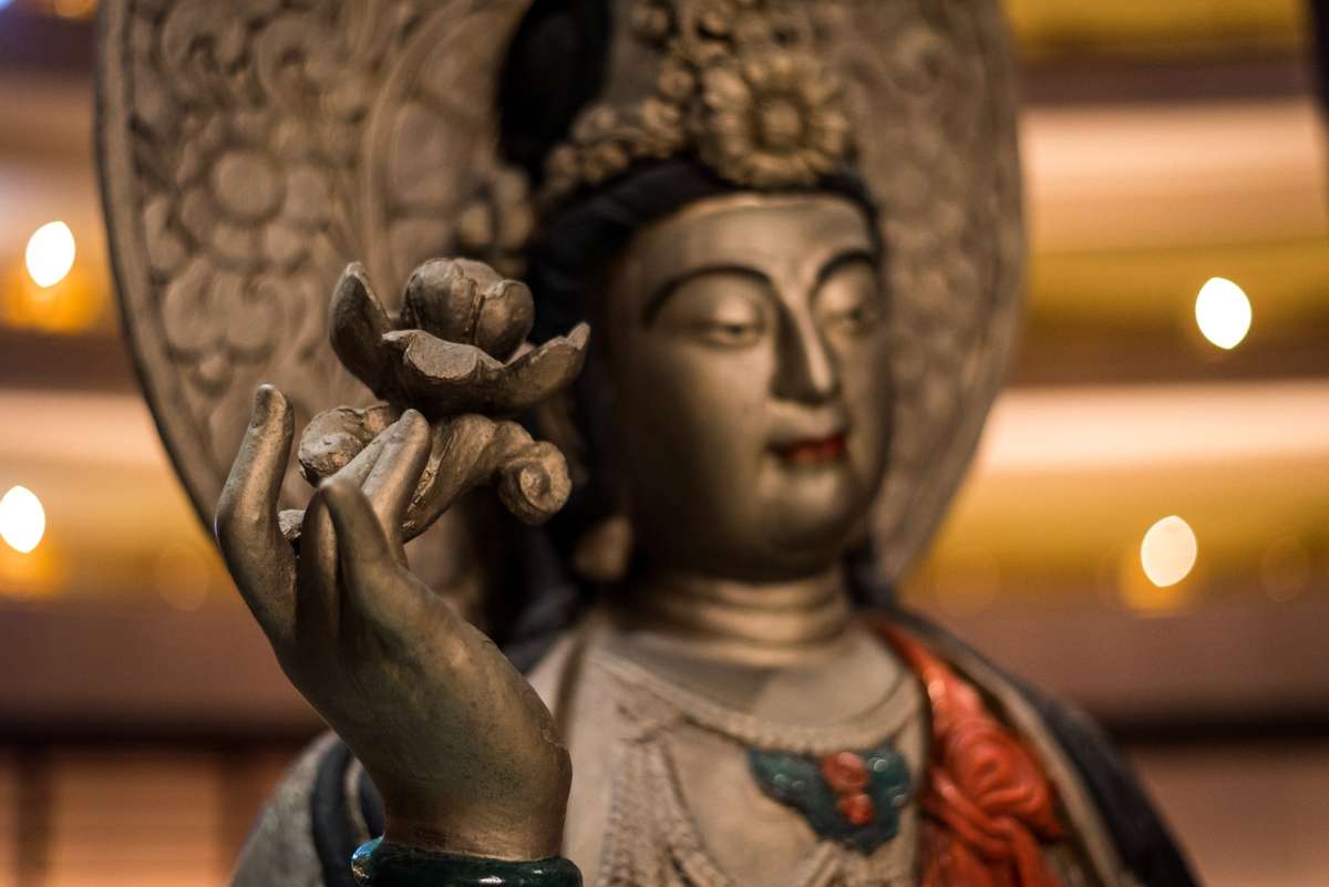 A golden Buddha statue with a serene expression holds a small offering in its hand against a blurred background of lights.