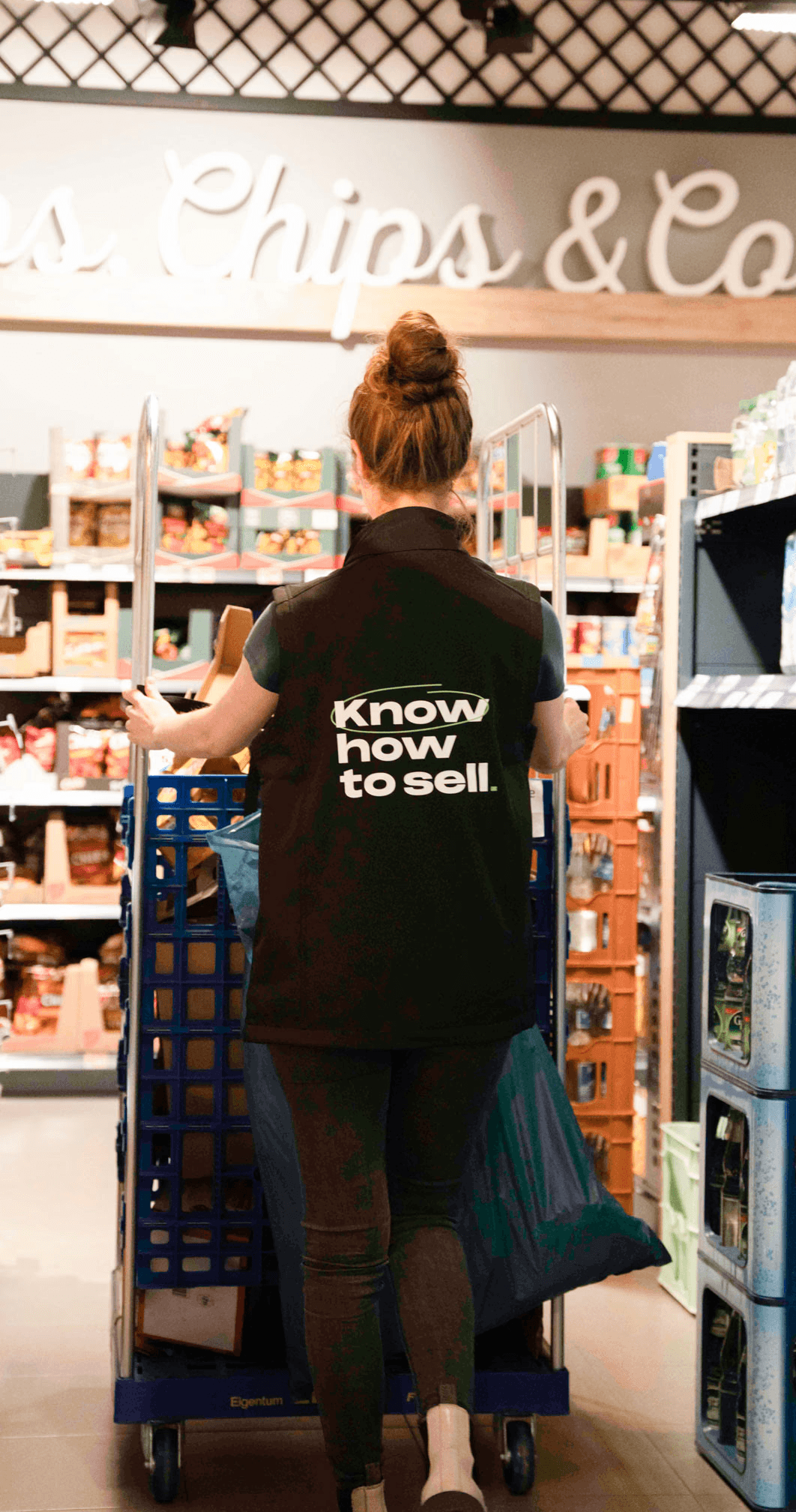 Mitarbeiterin im Lager mit Weste ‚Know how to sell‘ zwischen Regalen mit Waren. Zuverlässiger Lagerservice schafft Ordnung, sichert Warenverfügbarkeit und stärkt die Effizienz am POS.