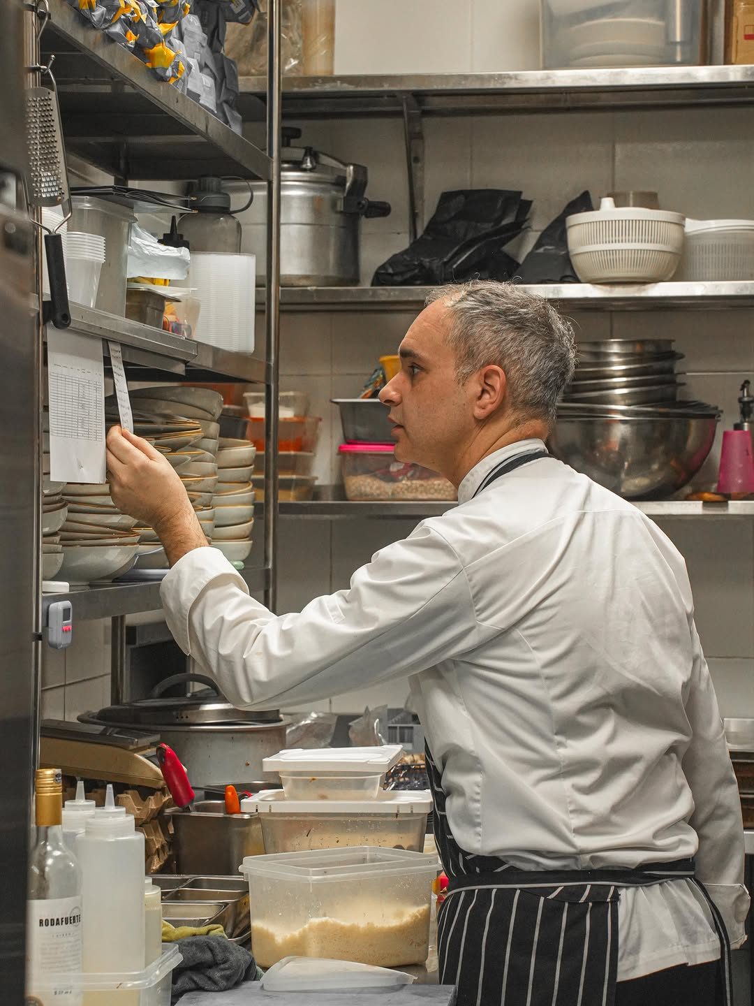 A chef in a kitchen reviewing a document on the wall, surrounded by various food preparation items and containers.