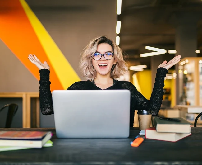 A delighted woman with glasses raises her hands in excitement in front of her laptop in a brightly lit, modern workspace.