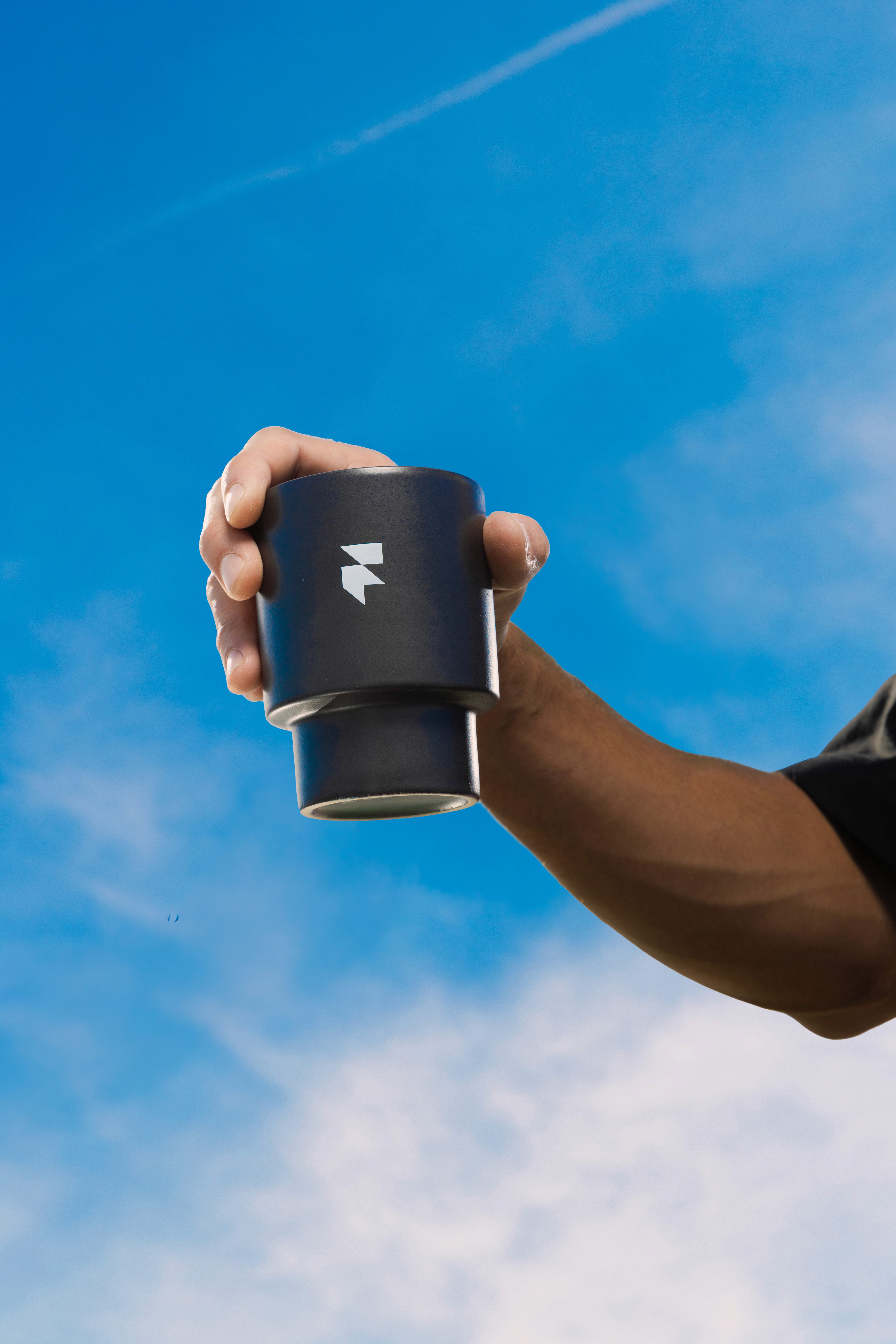 A person is holding a sleek, black travel mug with a geometric logo, set against a clear blue sky with wispy clouds, showcasing portable drinkware design.