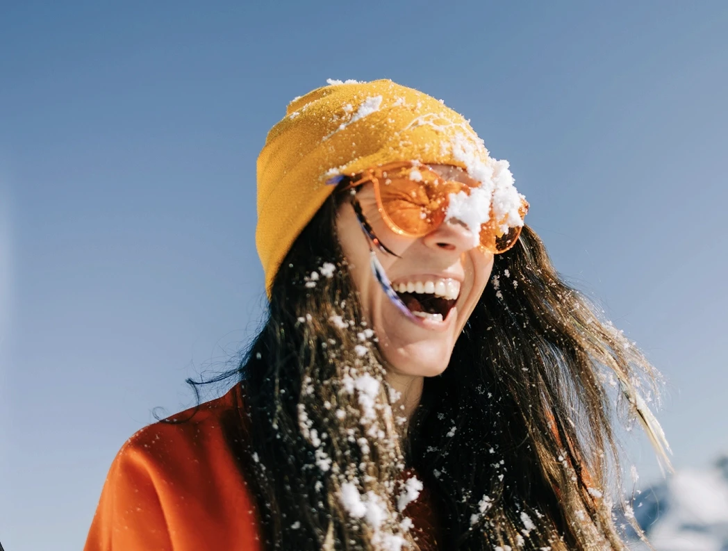 Laughing woman with snow-covered yellow beanie and orange sunglasses wearing red jacket outdoors against clear blue winter sky, joyful ski holiday lifestyle portrait
