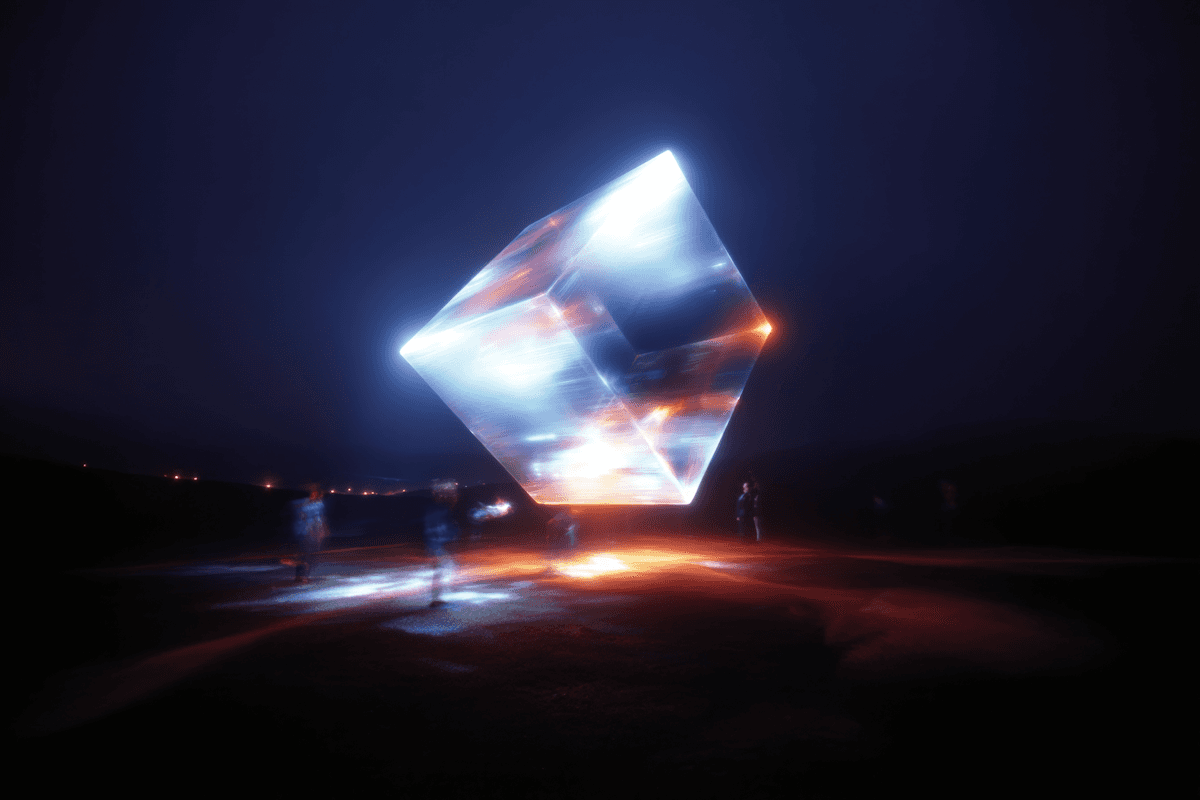 A glowing, floating cube illuminated in blue and orange hues at night, surrounded by blurred figures in a surreal outdoor setting.