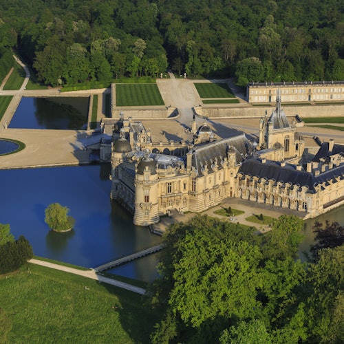 一座大型历史悠久的城堡航拍图，四周环绕着护城河、郁郁葱葱的花园和茂密的森林。