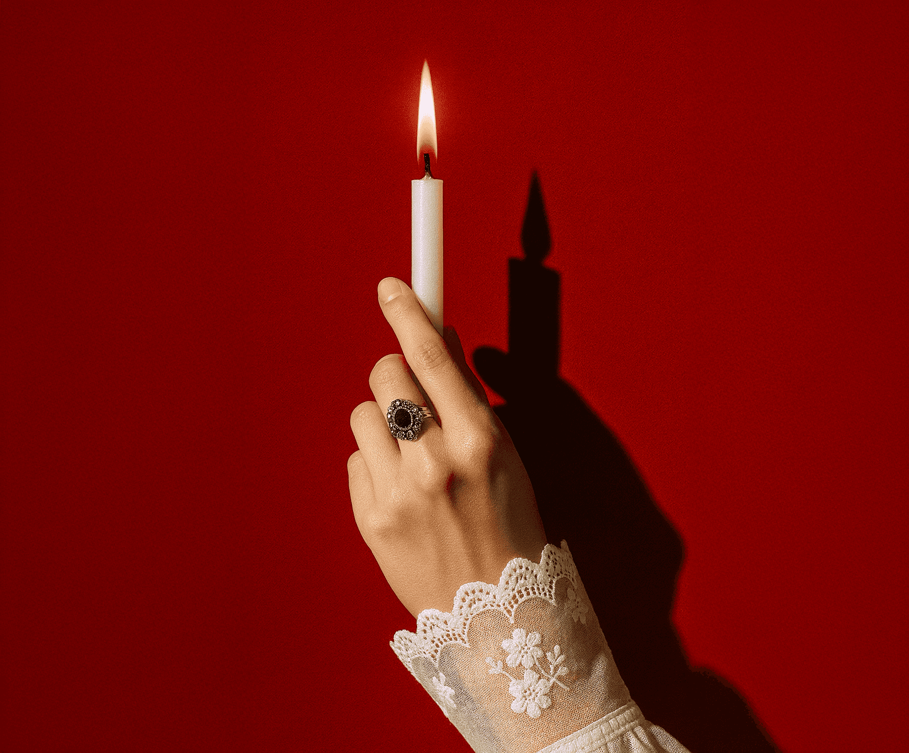Hand in lace sleeve holding a lit white candle against a red background, wearing a silver ring with a dark gemstone.