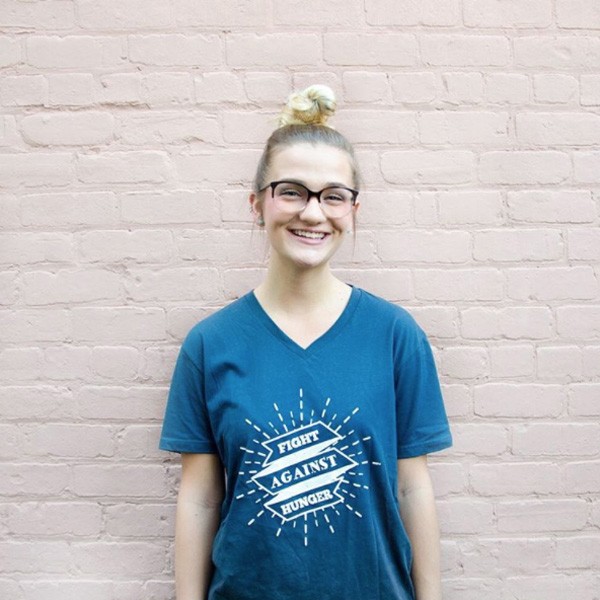 Person smiling wearing a "Fight Against Hunger" t-shirt, standing against a pink brick wall.