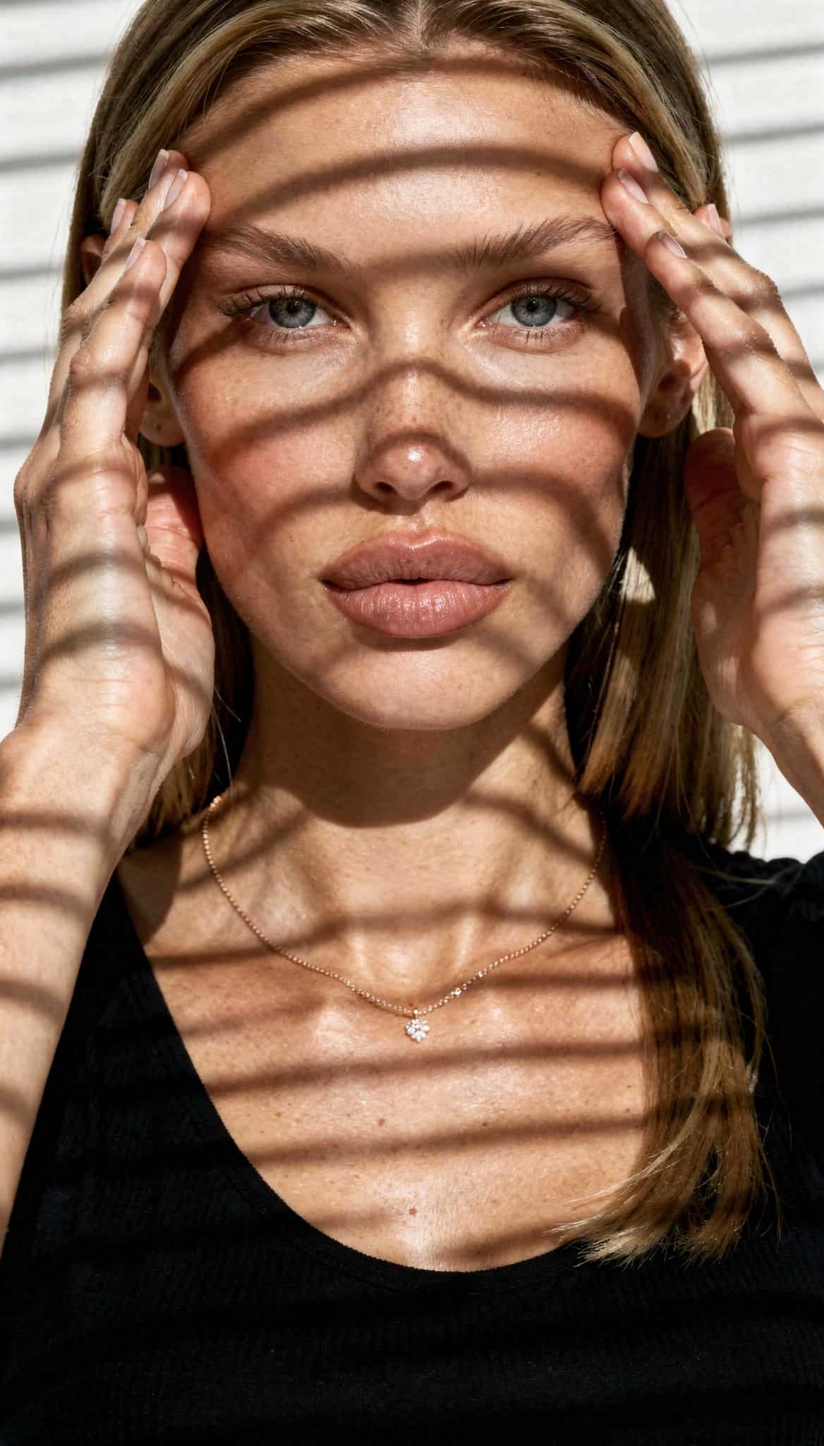 a young woman with blonde hair and blue eyes. She is looking directly at the camera, with her hands framing her face