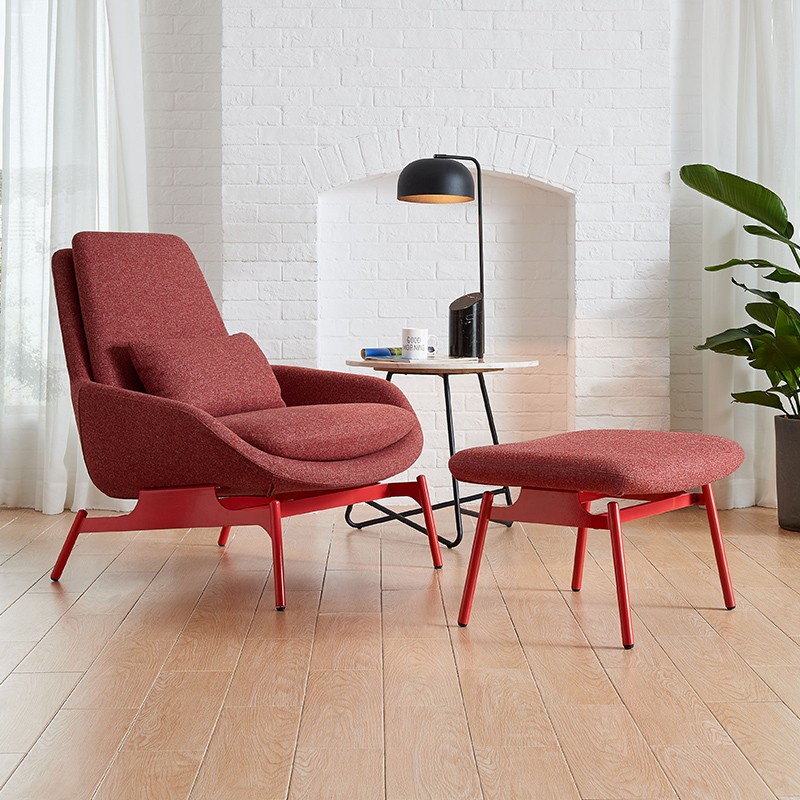 Mid-century modern red lounge chair and matching ottoman set with a textured fabric finish and red sculptural metal legs, positioned in a bright room with a white brick fireplace, marble side table, and indoor bird of paradise plant.