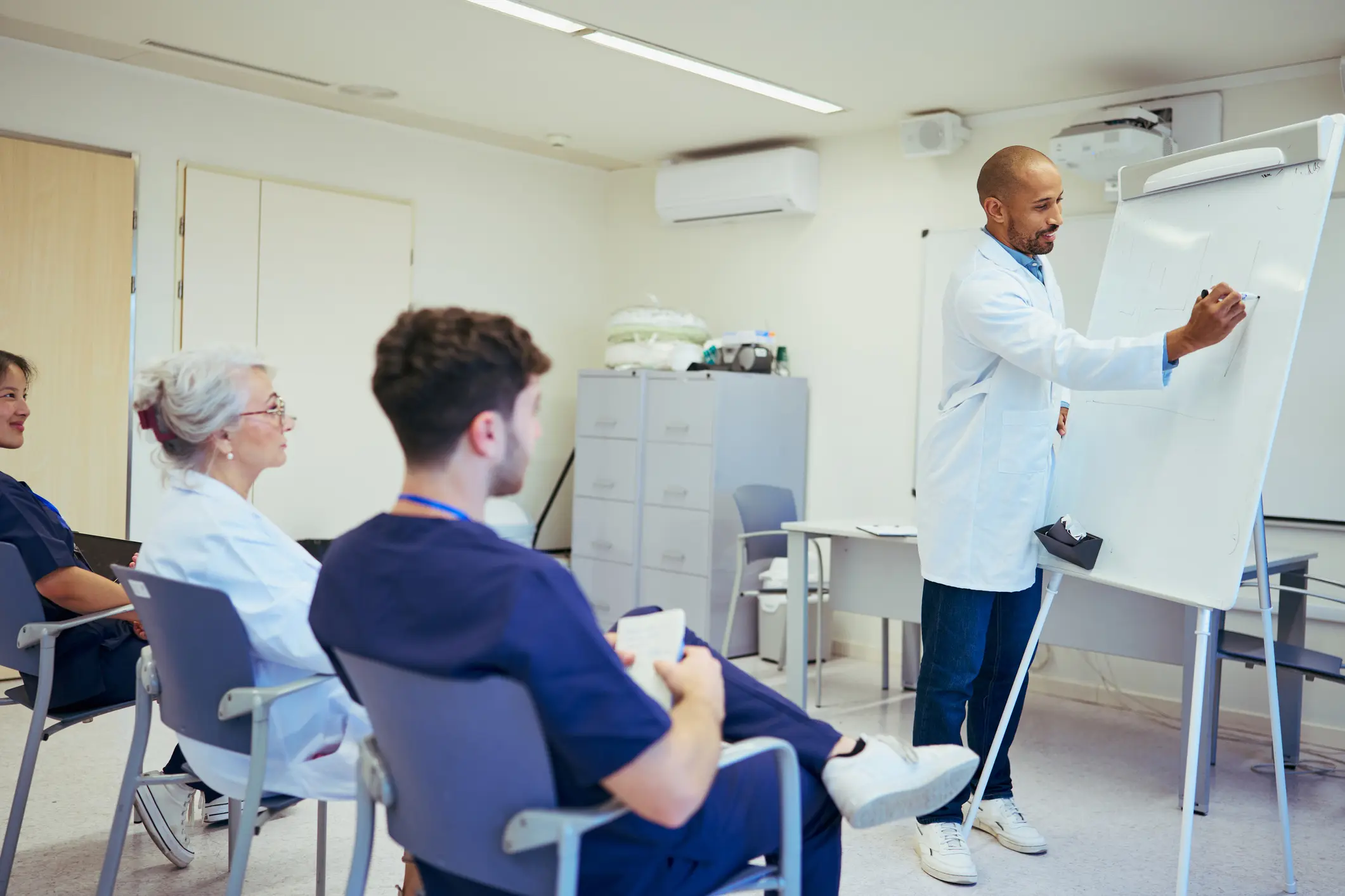 Healthcare professional in white coat presenting at a flipchart to a seated team of doctors and nurses, illustrating coaching and capability building in upstream leadership