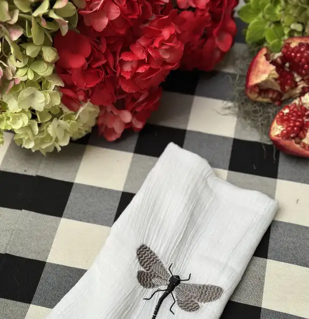 Detalle de mesa con servilleta bordada con libélula sobre mantel a cuadros, flores rojas y granada abierta para composición otoñal elegante.