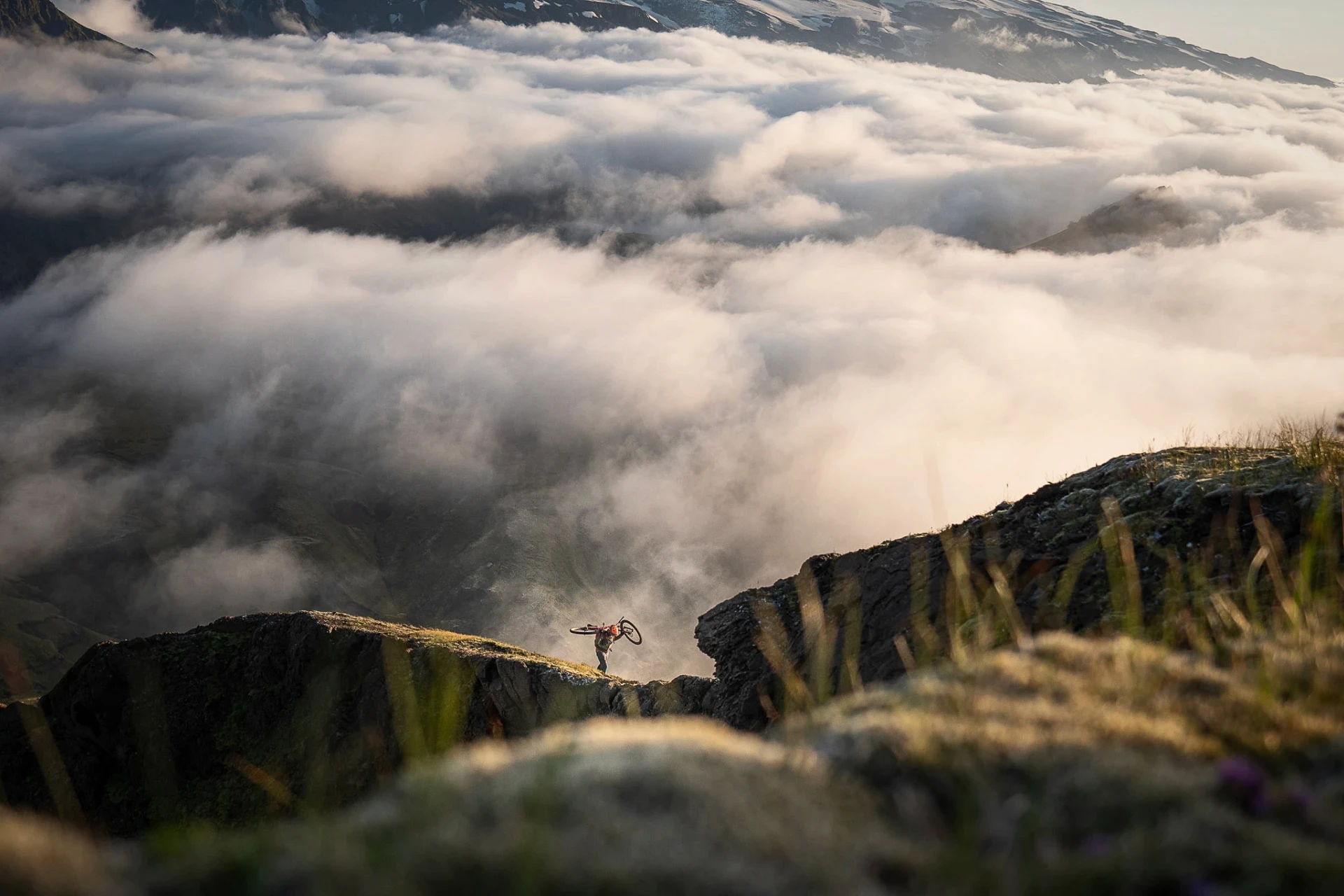 Person carrying a bicycle on a mountain ridge above low clouds.