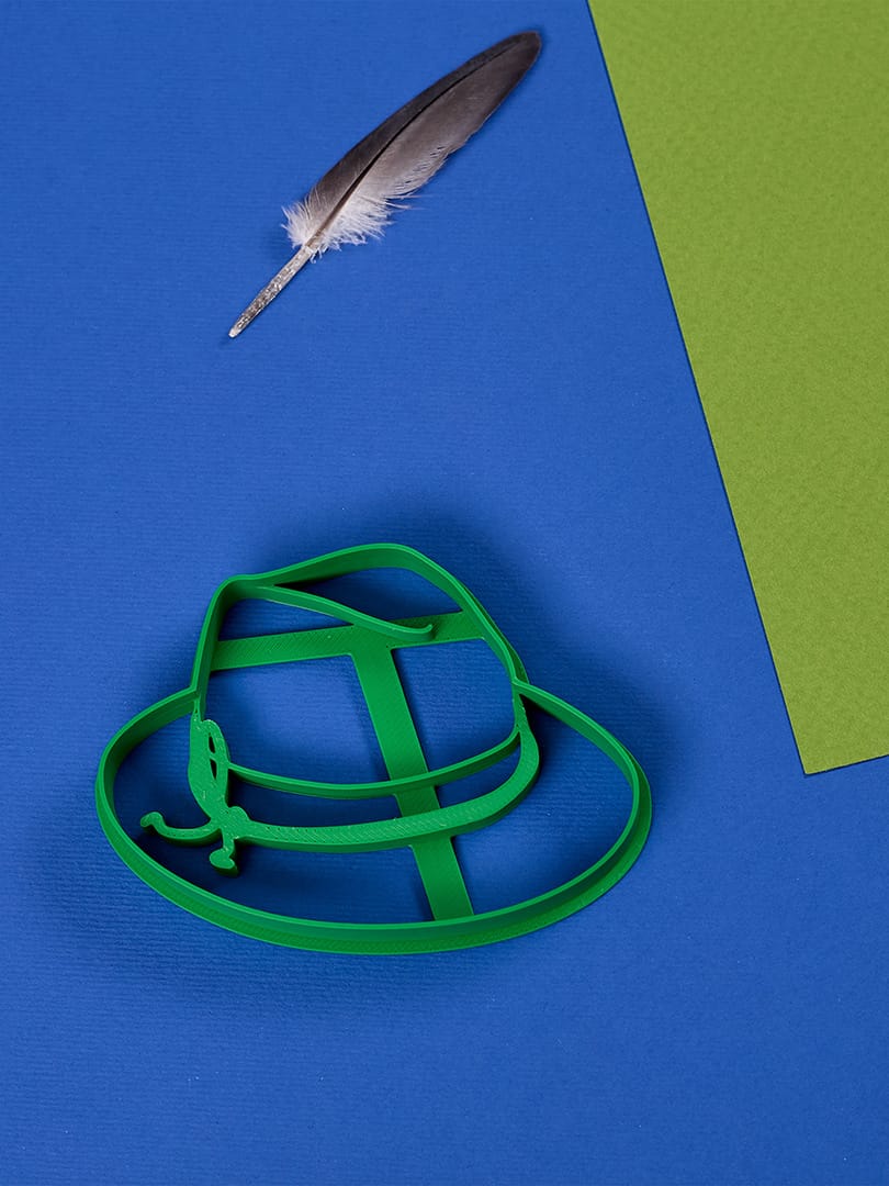 Zielona foremka do ciastek w kształcie kapelusza od Biocraftlab, na tle żywej niebiesko-zielonej scenerii, z jednym piórem podkreślającym kreatywną i zabawną estetykę.