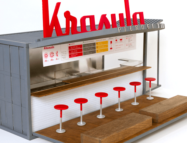 A gray and white model of a Krasila Pierogi Bar with red lettering spelling “Krasila” above the counter and a menu board listing food and drink items. Six red bar stools are arranged along the counter. The model is positioned against a white background.