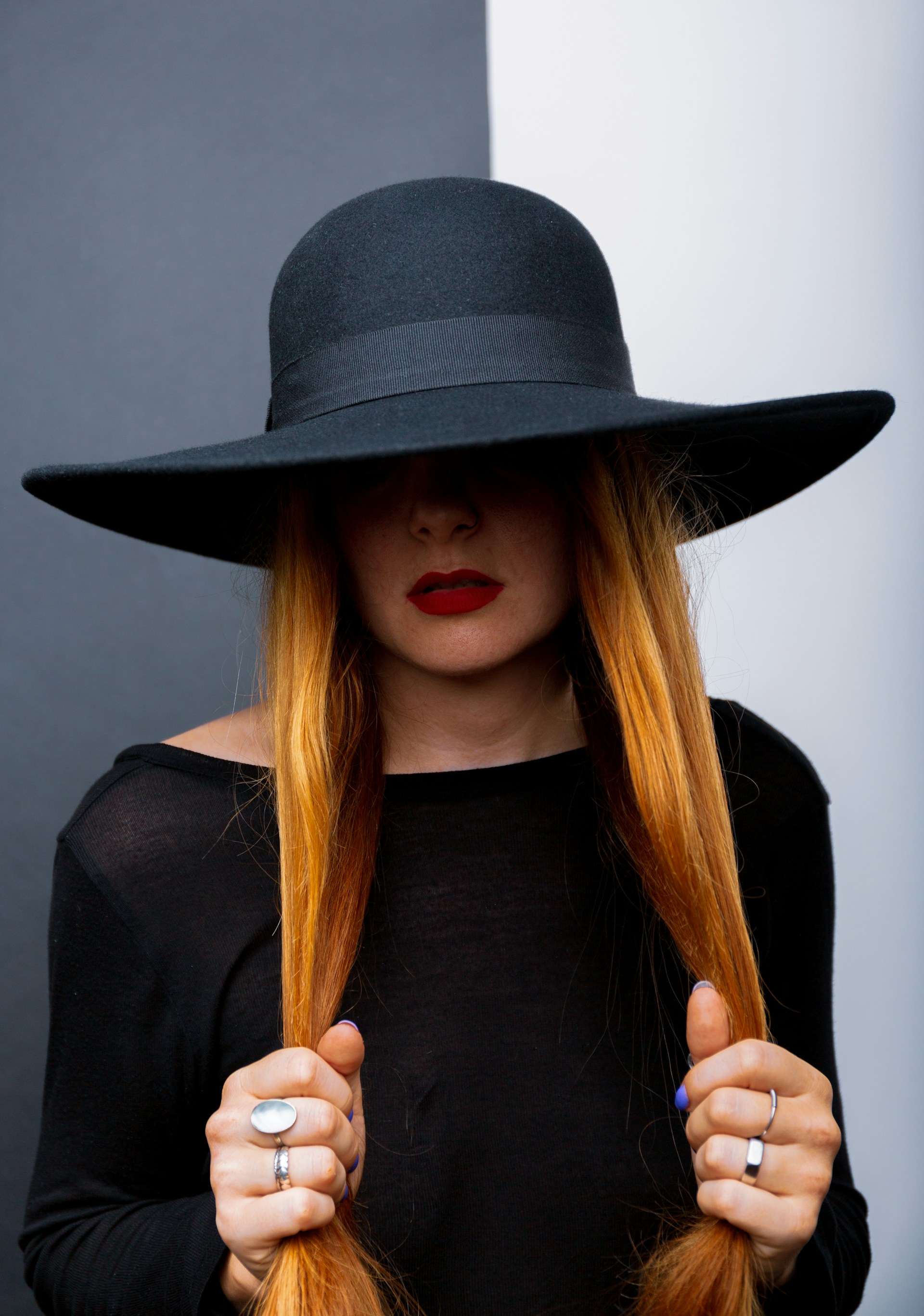 A woman in a black wide-brimmed hat and black top holding two sections of her long ginger hair. Her eyes are obscured by the hat's shadow, highlighting her red lipstick and various silver rings.