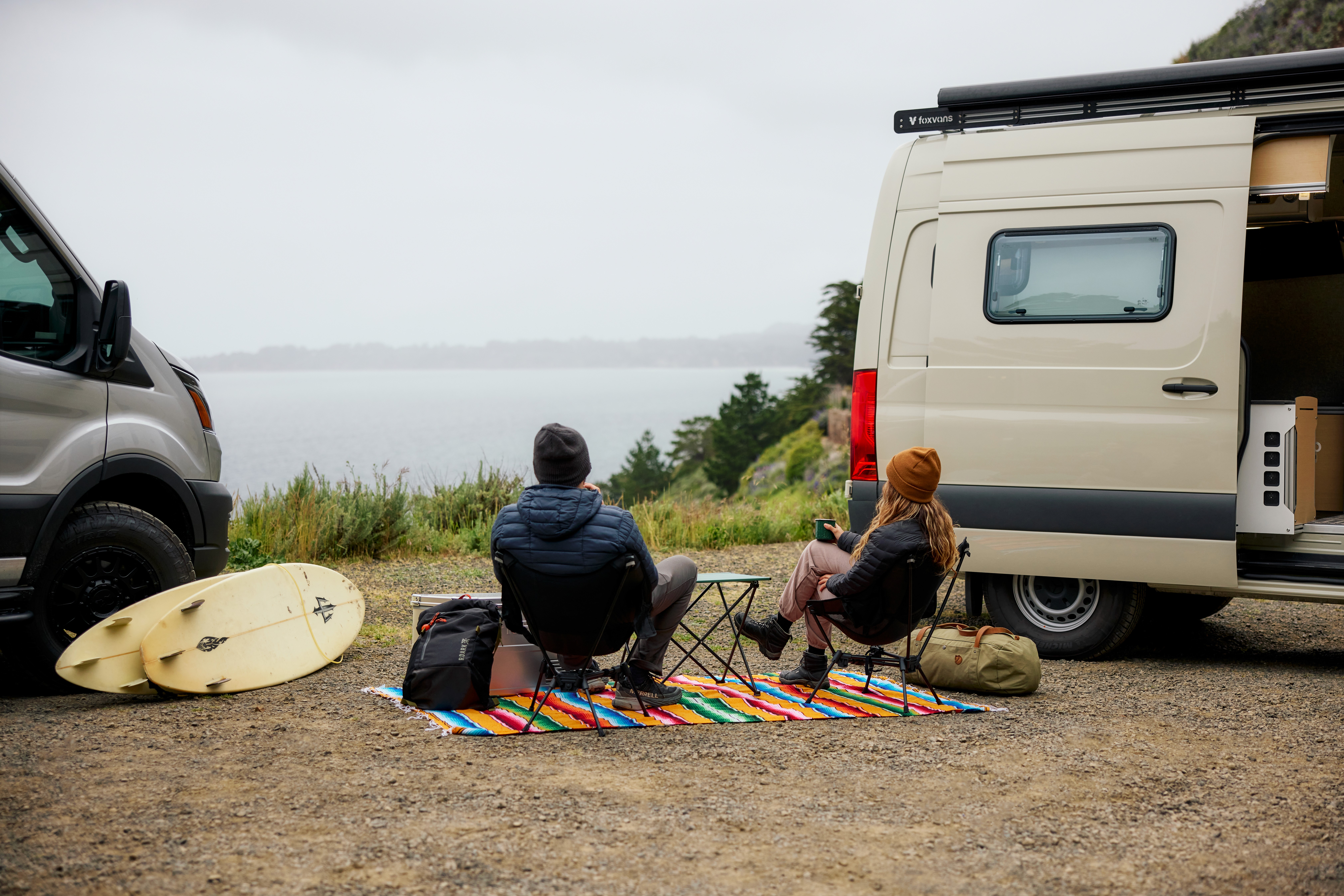 Two people sit on the ground near a camper van, with surfboards nearby, overlooking a coastal view.