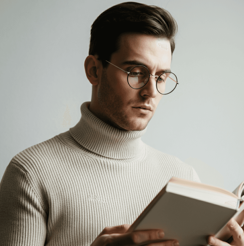 A man wearing round glasses and a cream-colored turtleneck sweater is intently reading a book against a plain gray background.