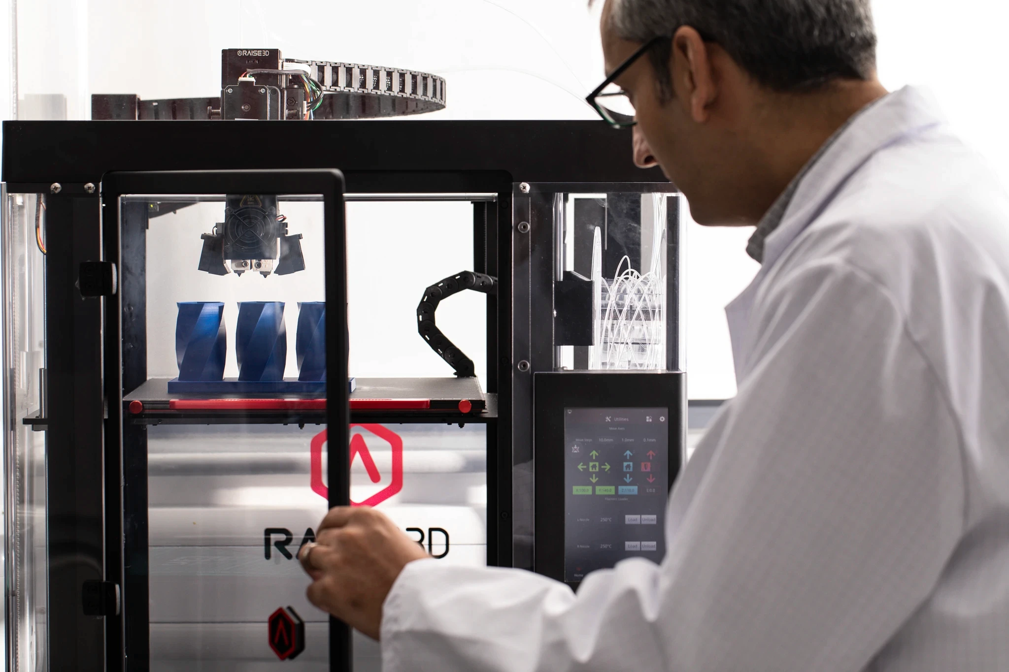 Scientist in a lab coat and safety glasses operating a 3D printer producing blue printed parts.