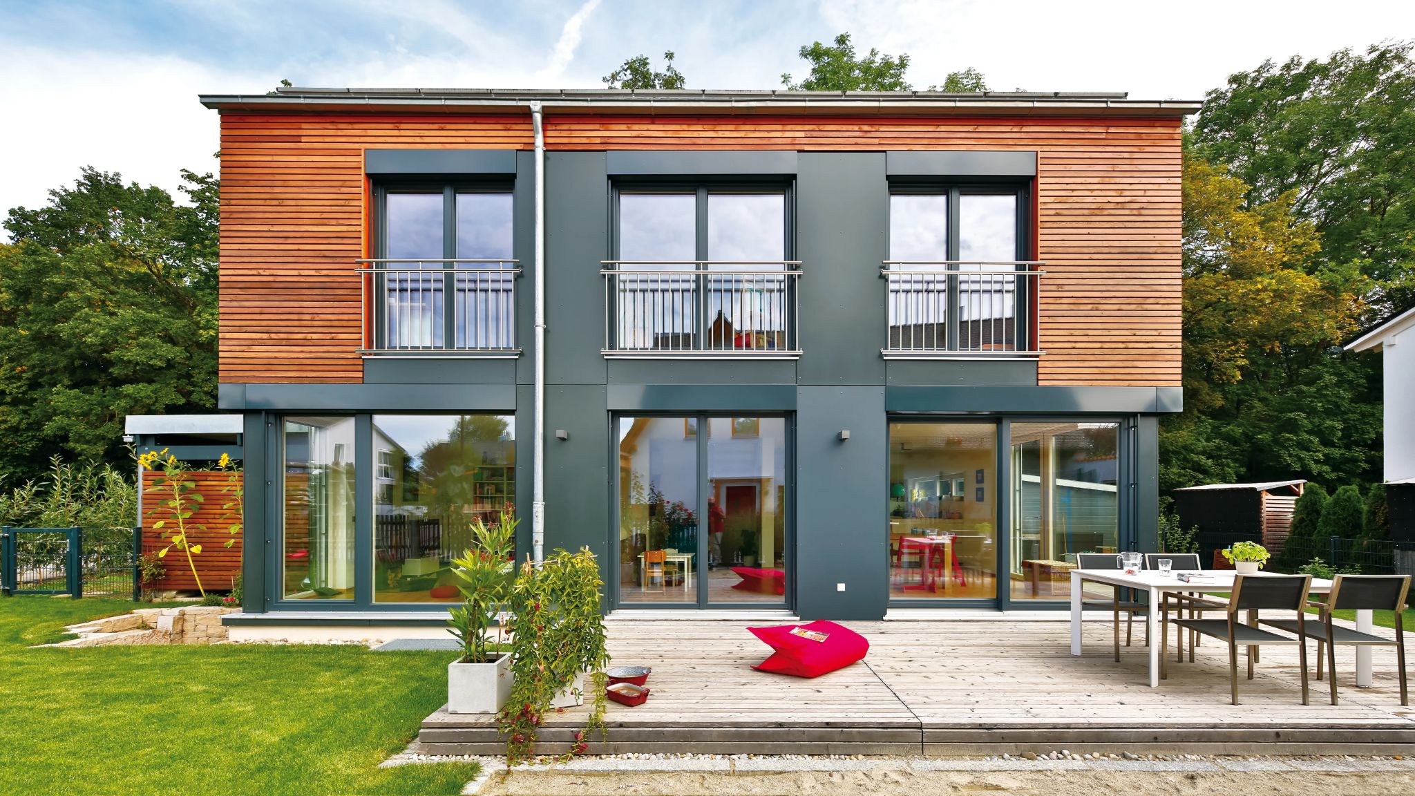 Modernes Einfamilienhaus mit Holzfassade im Obergeschoss, dunkler Verkleidung im Erdgeschoss und großer Glasfront, Terrasse mit Gartenmöbeln.