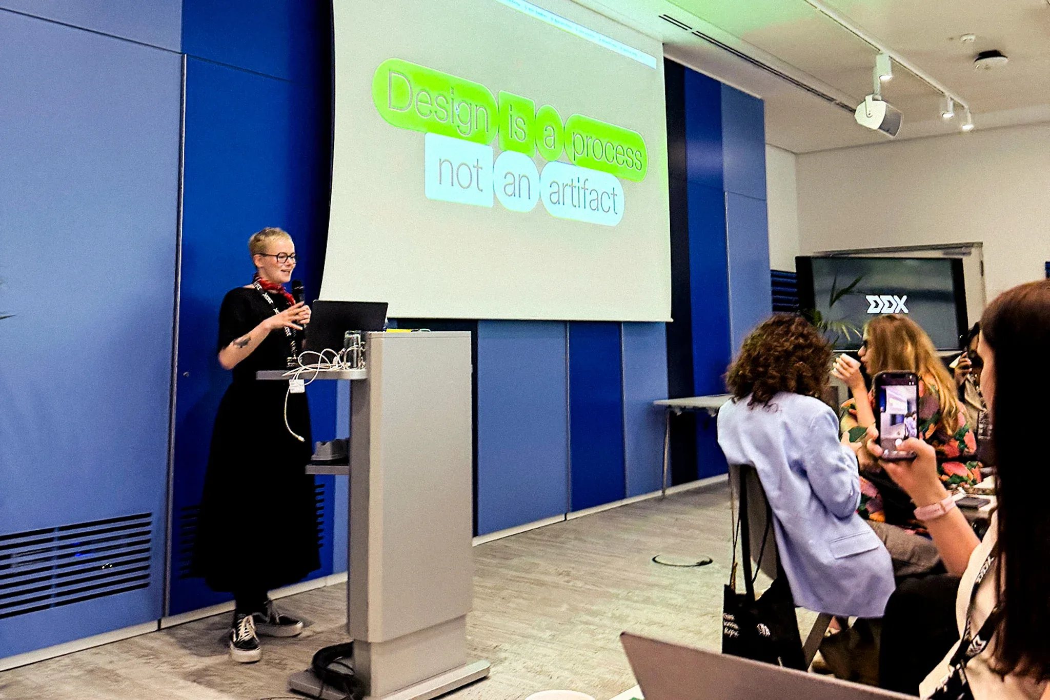 Woman speaking at the DDX conference with presentation showing that design is a process not an artifact