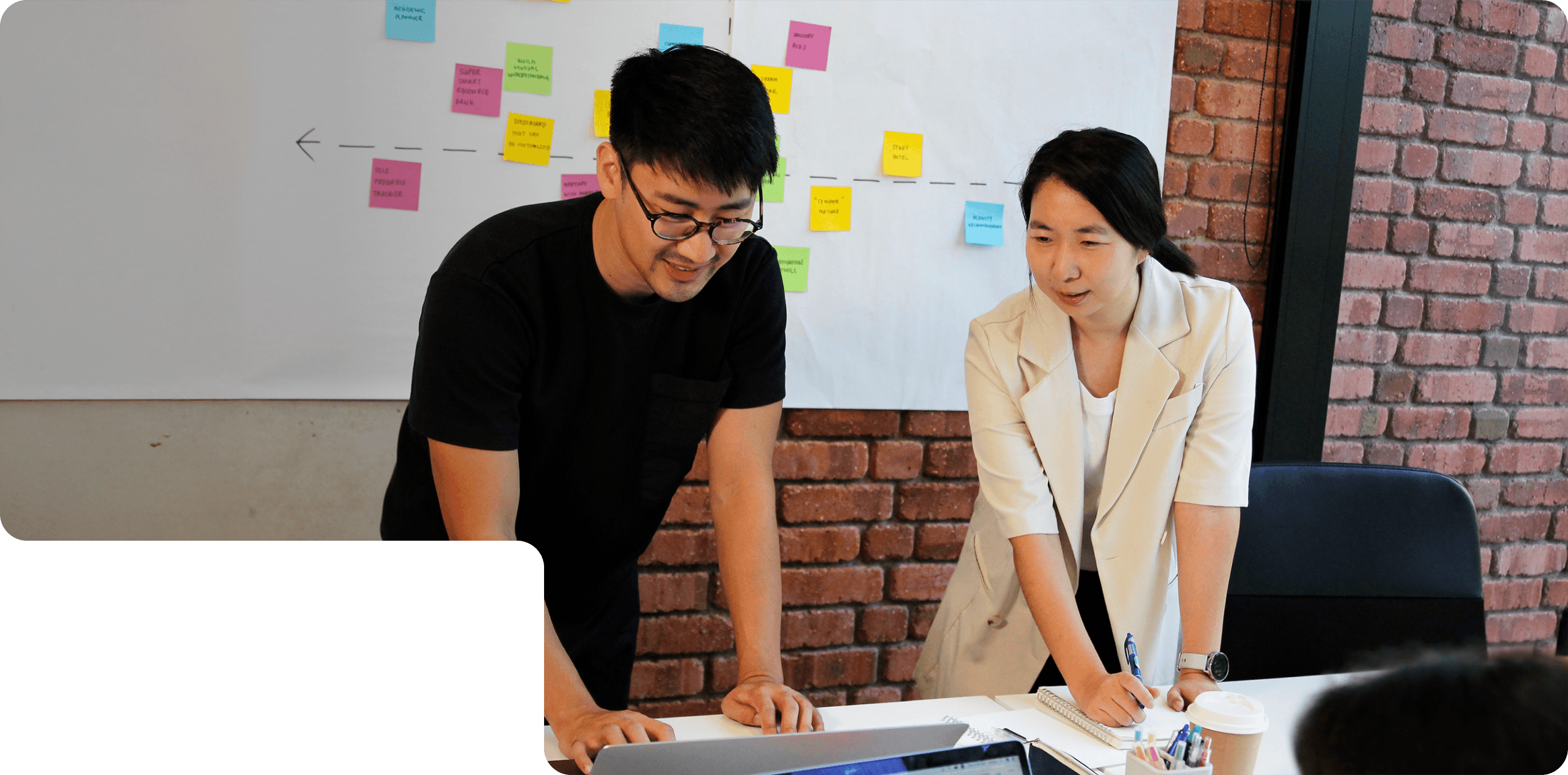 Two members of 55 Minutes collaborating in a modern workspace, discussing ideas in front of a laptop and a board with sticky notes.