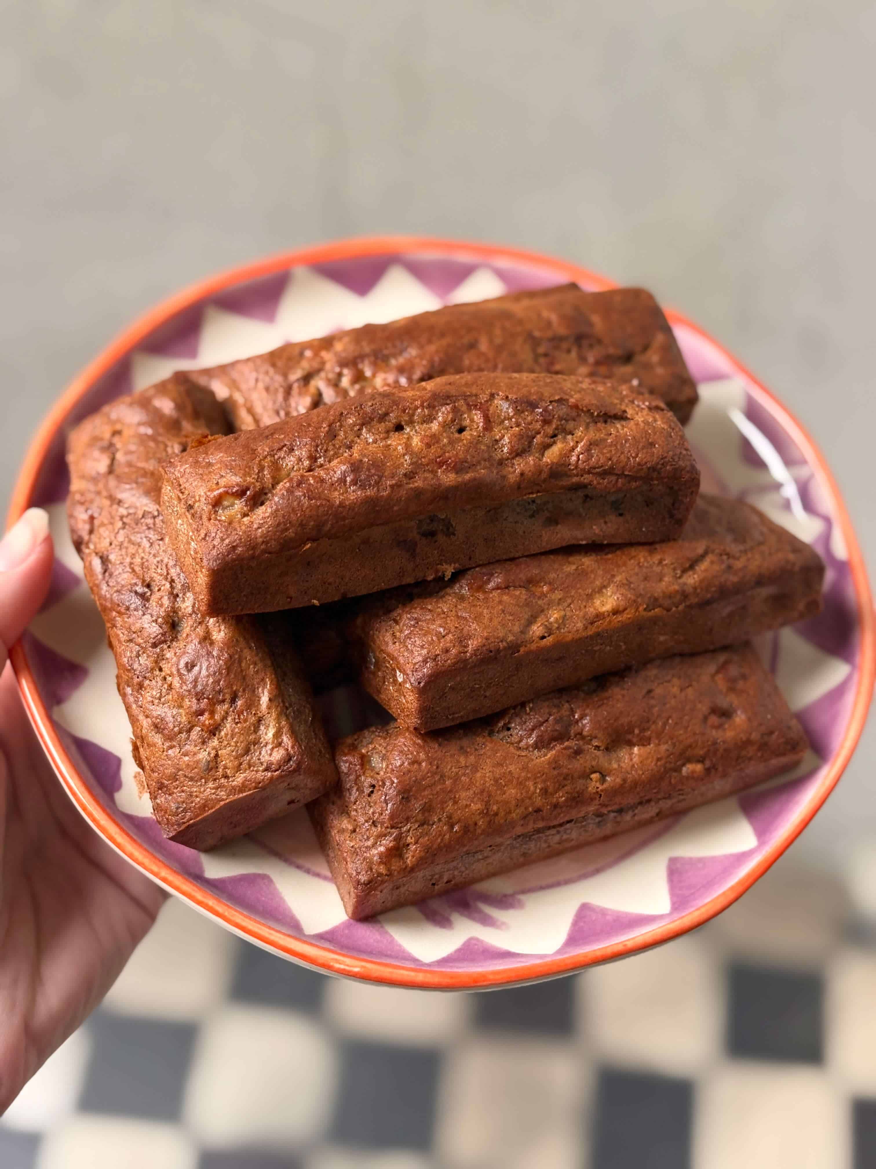Mini walnut banana bread loaves stacked on a patterned plate, showing a moist texture with visible banana and walnut pieces.