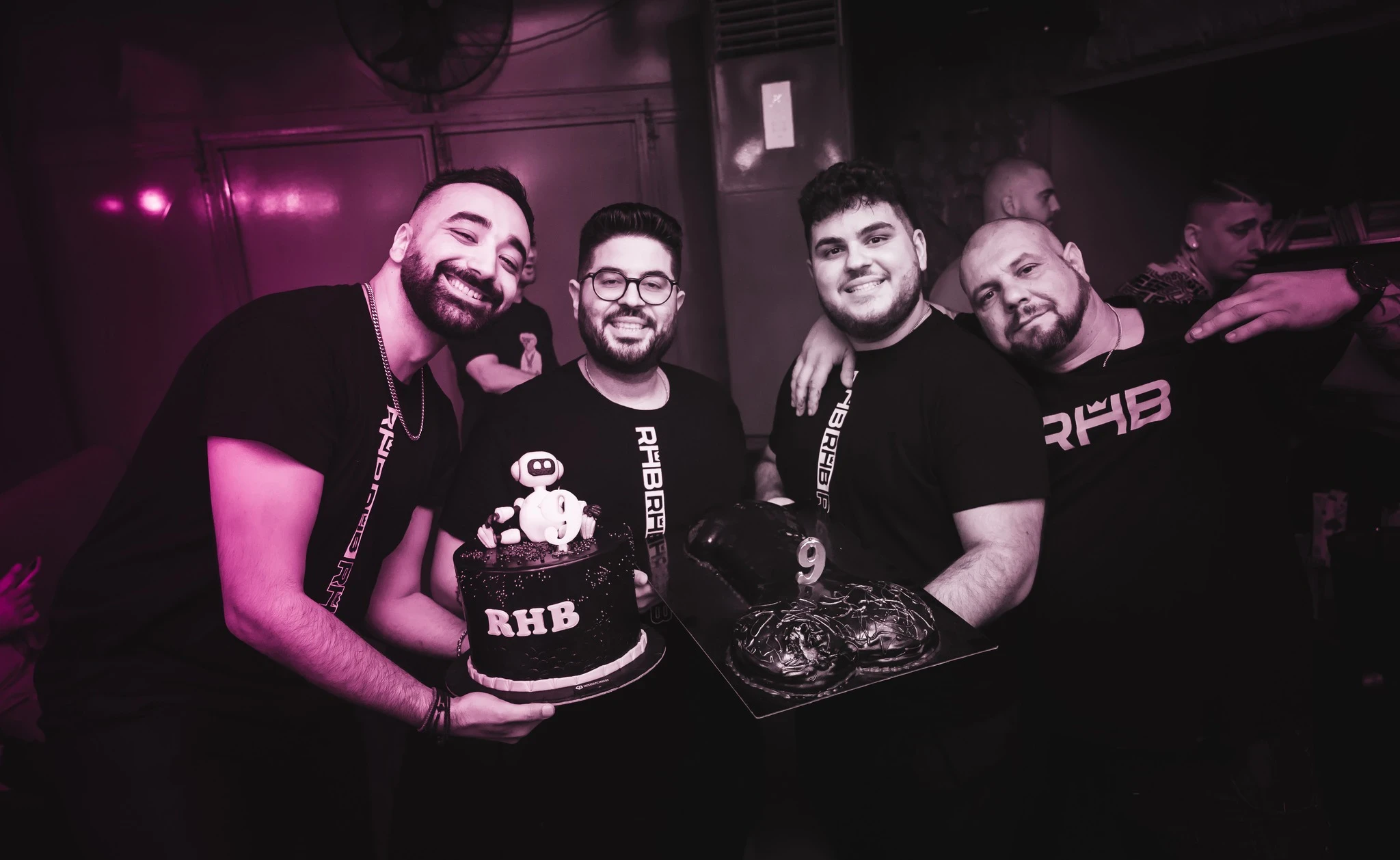 Four men in matching black "RHB" t-shirts smile and pose for the camera. Two of them hold cakes: one is a round "RHB" cake with a robot topper, and the other is a chocolate cake with the number 9.