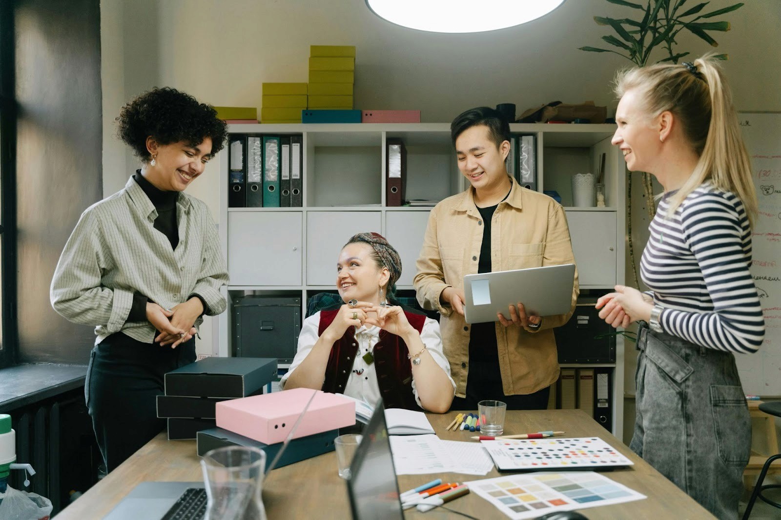 For people in an office space. There is three women and one man, they are all laughing. There is laptops books and boxes in front of them on the table.