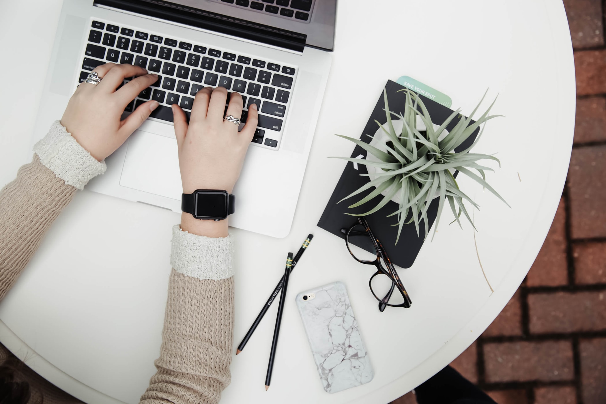 A person's hands are typing on a silver laptop, surrounded by a minimalist workspace with a plant, glasses and a phone.