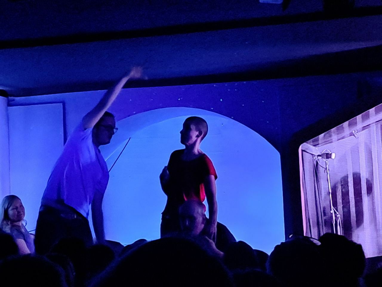 Zwei Schauspieler auf der Bühne eines Improtheaters, beleuchtet von blauem Licht. Eine Person in einem weißen T-Shirt hebt den Arm, während die andere Person, im roten T-Shirt, sie beobachtet. Eine Zuschauerin sitzt im Vordergrund.