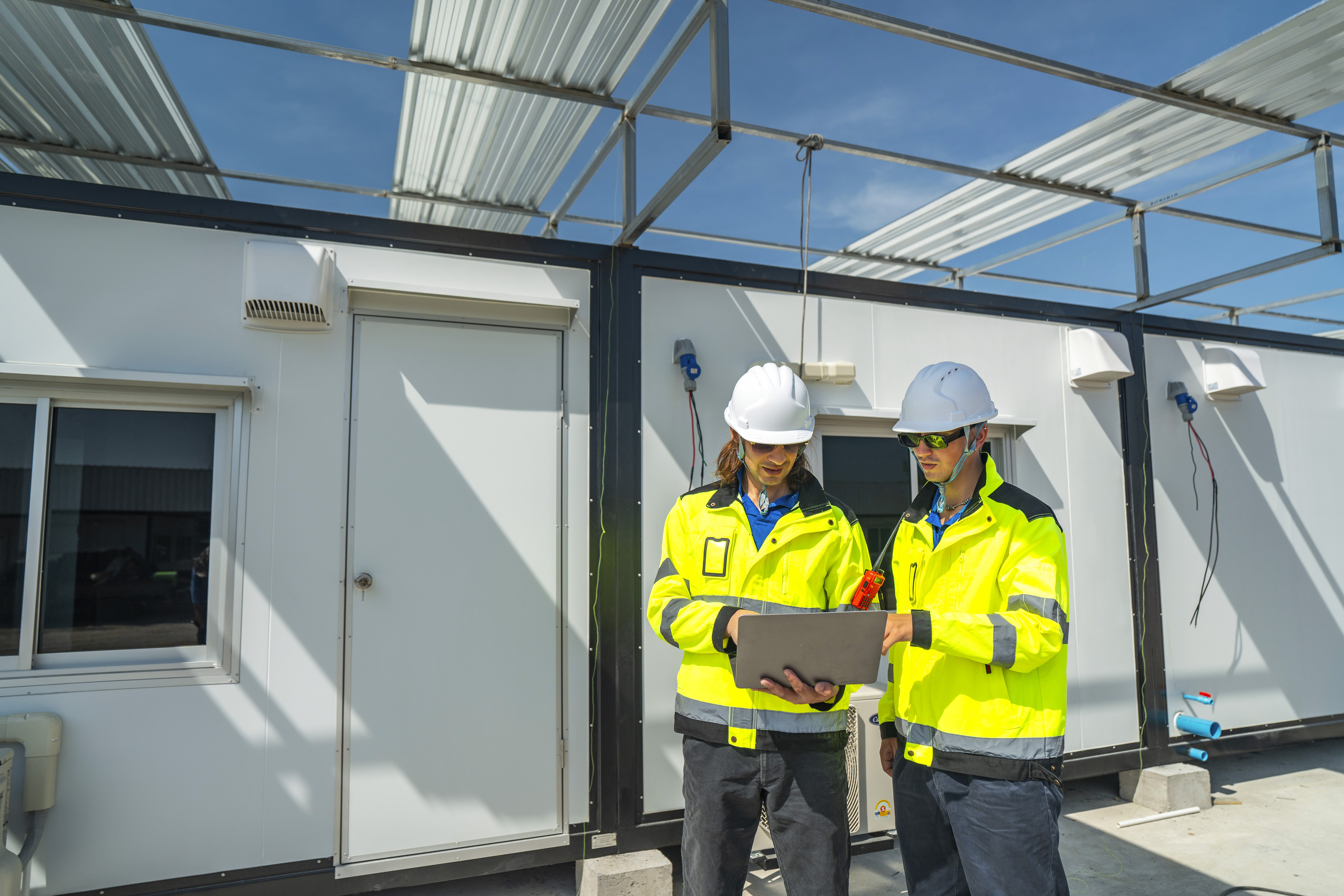 Two engineers in white helmets and yellow jackets in front of a building discussing construction plans.