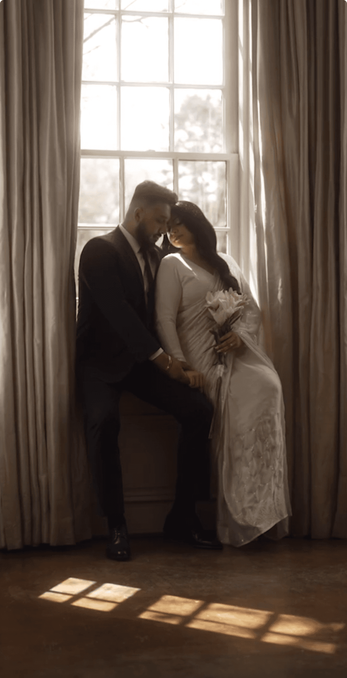 Bride in a white dress holding a bouquet sits beside the groom in a black suit on a window sill, captured in an intimate wedding moment.