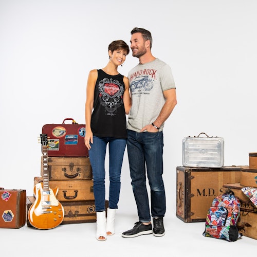 Sepasang pasangan yang tersenyum berdiri di samping koper vintage, gitar, dan ransel berwarna-warni dengan latar belakang putih.