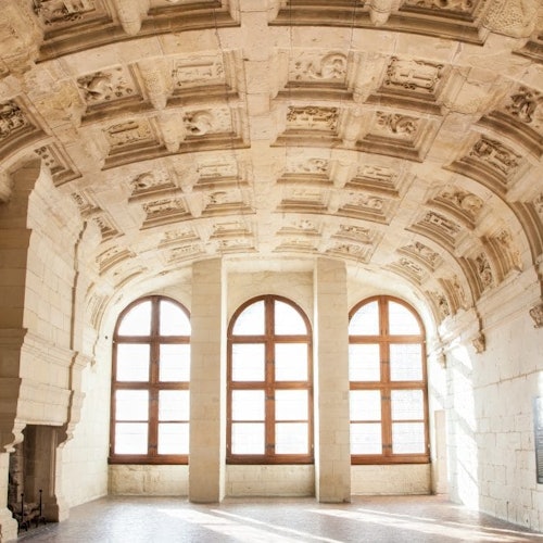 Un plafond voûté et orné avec des sculptures minutieuses, trois grandes fenêtres cintrées, et la lumière du soleil inondant une salle vide au sol en pierre.